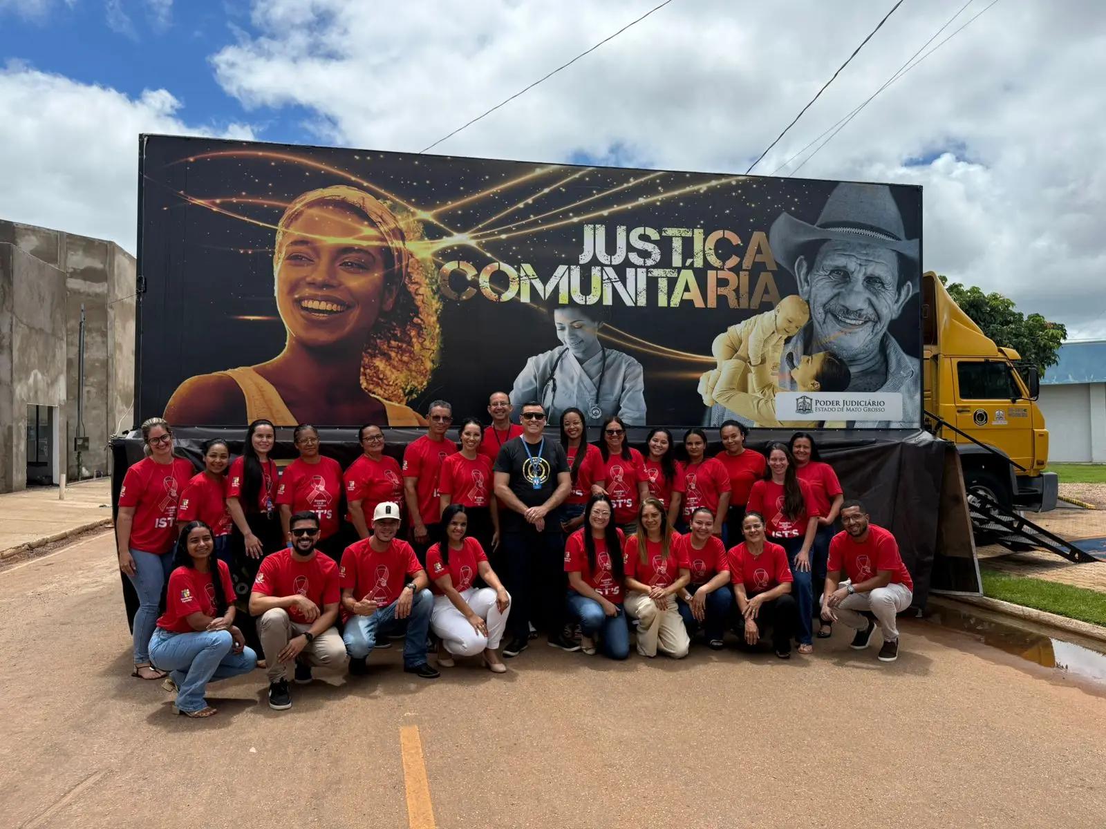 Equipe do programa Justiça Comunitária reunida em frente ao caminhão itinerante. Cerca de 25 pessoas vestem camisetas vermelhas com a identificação do projeto e posam para foto em grupo. Ao fundo, o veículo amarelo exibe painel com a inscrição "Justiça Comunitária".
