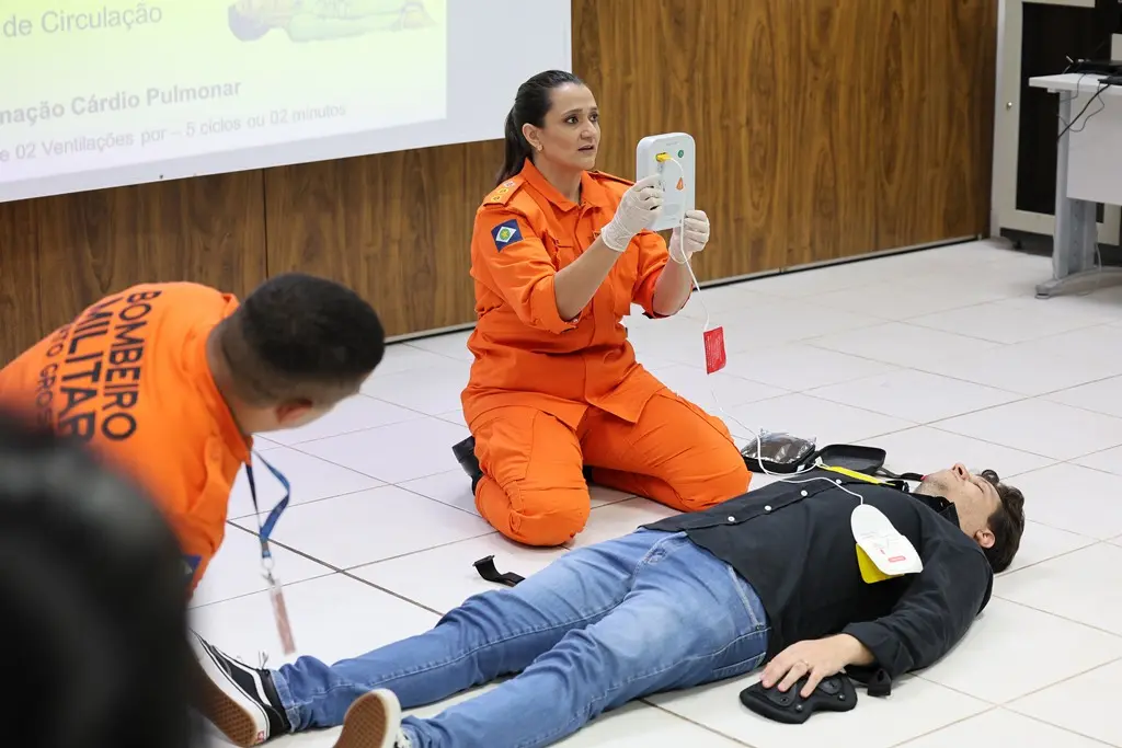 Foto horizontal que mostra uma aula prática sobre uso de desfibrilador. A instrutora, tenente-coronel bombeiro militar Aline Regina Novacki Nunes, está agachada ao chão, ao lado de um aluno, que está deitado ao solo. Ela mostra o desfibrilador para os alunos. Ao lado, há outro bombeiro instrutor.