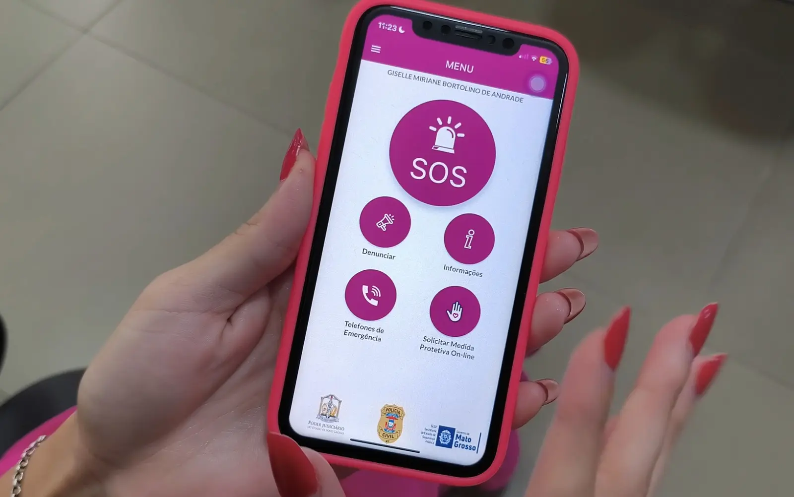 Foto na horizontal que mostra as mãos de uma mulher manuseando um celular cuja tela está aberta no aplicativo SOS Mulher MT, que tem entre suas funções solicitar medida protetiva.