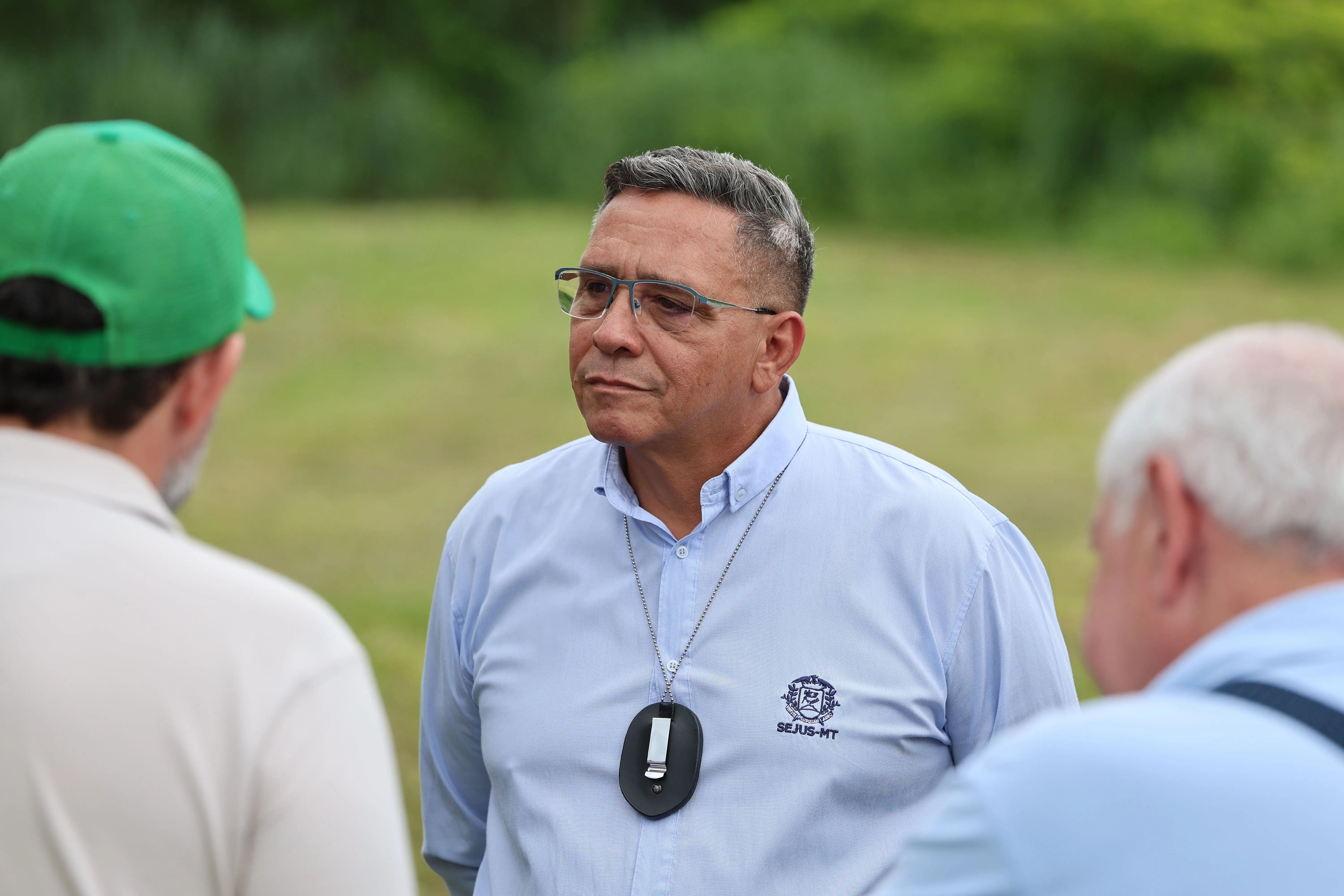 Close-up de um homem com óculos e camisa social azul clara. Ele usa um crachá de identificação pendurado no pescoço. Ele está conversando com duas outras pessoas.
