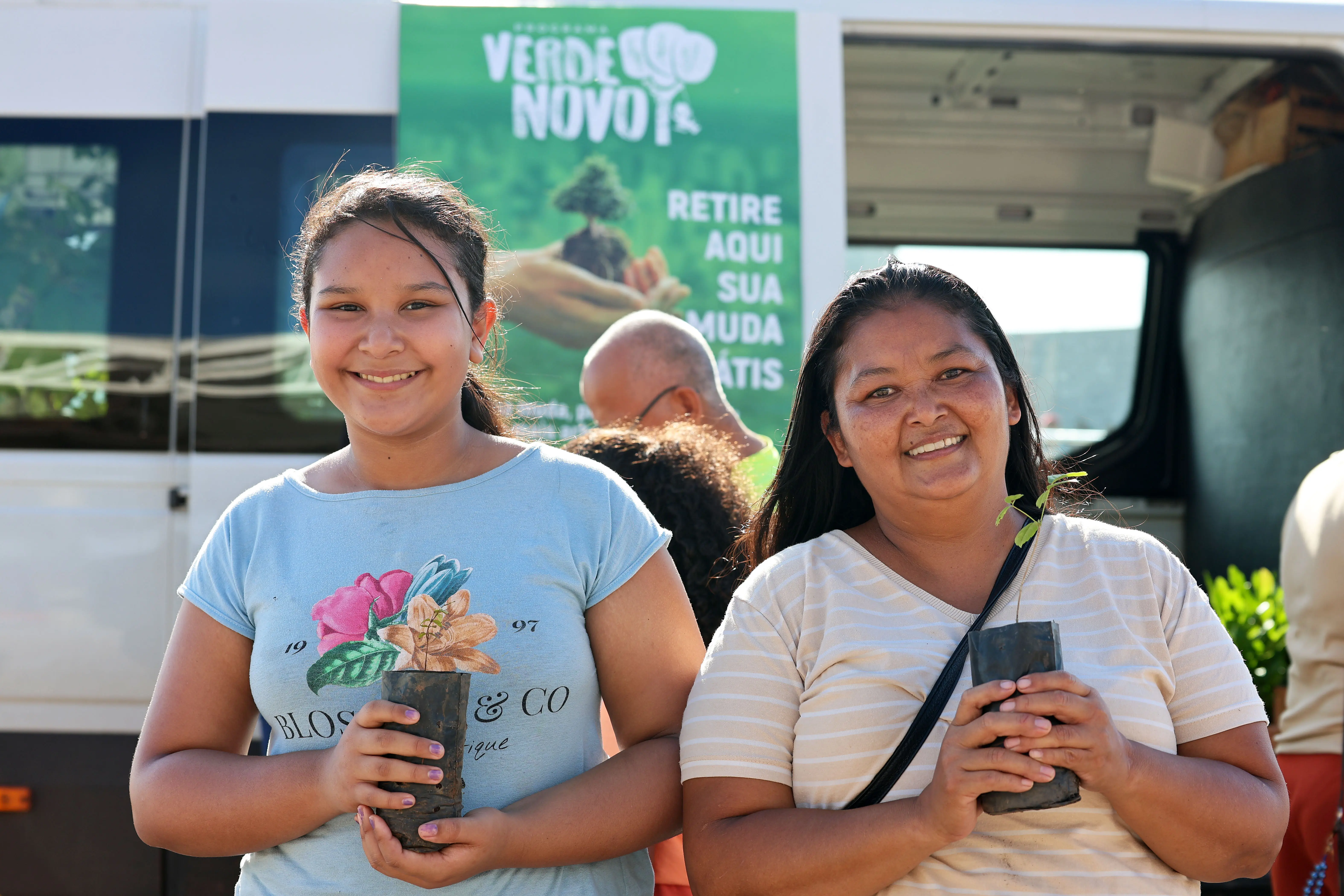 Mulher e jovem sorriem segurando mudas de árvores. Ao fundo, uma van do projeto 