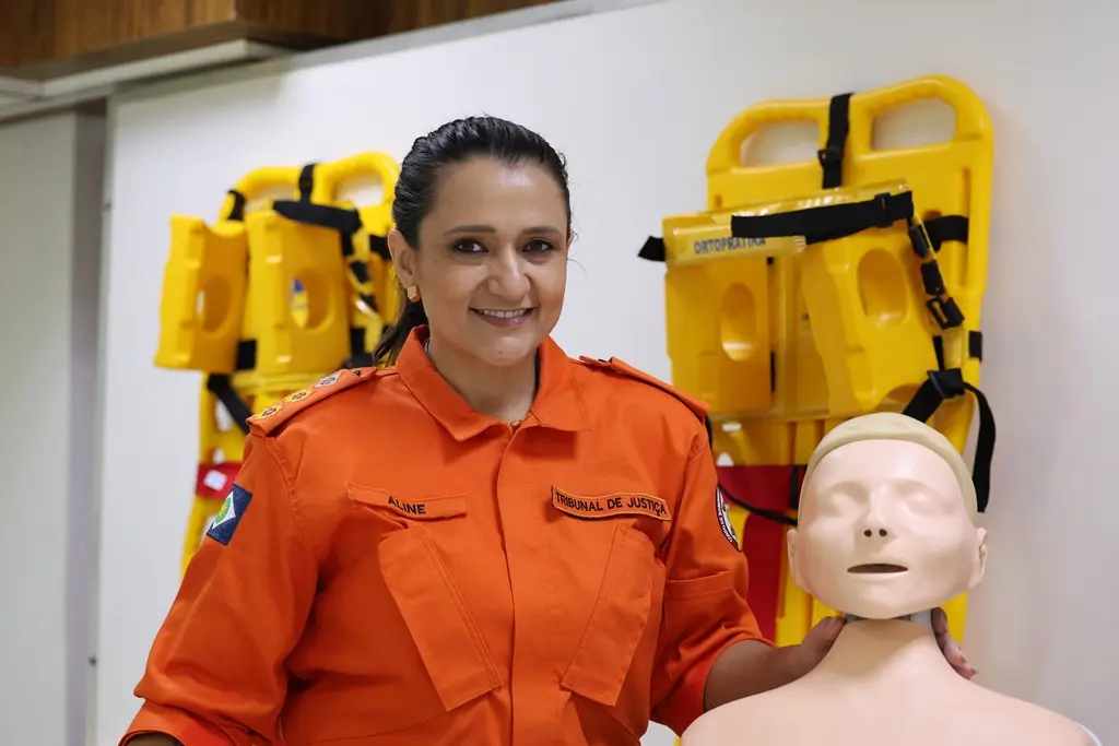 Foto horizontal da tenente-coronel bombeiro militar Aline Novacki posando sorrindo e segurando um boneco utilizado nas aulas práticas de primeiros socorros. Ela é uma mulher de pele clara, olhos e cabelos castanhos, cabelo preso, fardada. Atrás dela, há duas macas amarelas encostadas na parede.