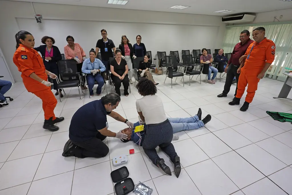 Foto horizontal, em plano aberto, que mostra uma sala de aula, onde três alunos simulam uma reanimação cardiopulmonar com uso de desfibrilador e os demais alunos e os instrutores, um bombeiro e uma bombeira, estão em volta, observando. 