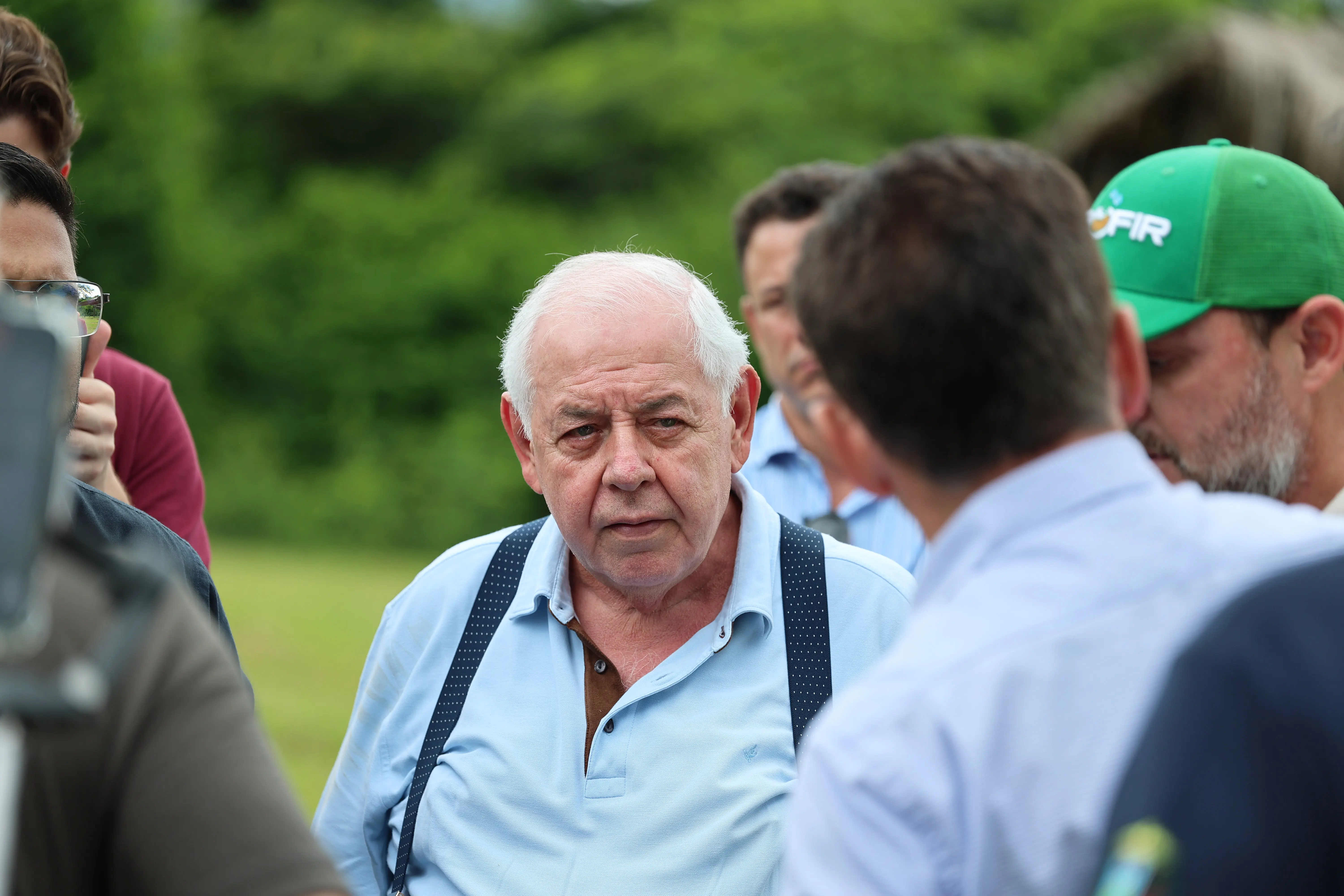 Close-up de um senhor, branco, com cabelos brancos e curtos, vestindo uma camisa polo azul clara e suspensórios. Ele está olhando diretamente para a câmera, em pé em um campo gramado.
