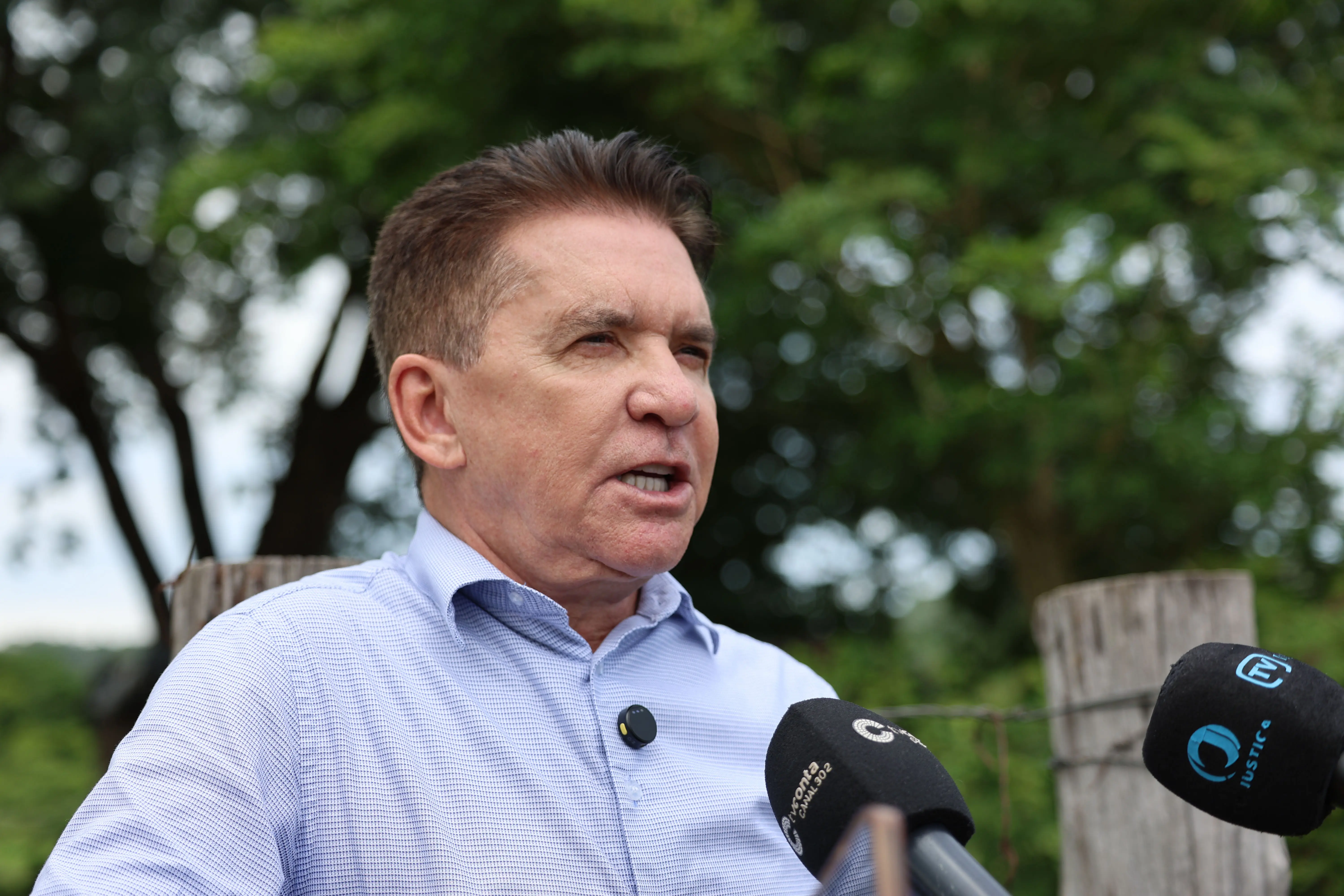 Close-up de um senhor, com camisa social azul clara, falando. Há dois microfones à sua frente. Ele está ao ar livre, com árvores e uma cerca de madeira ao fundo.