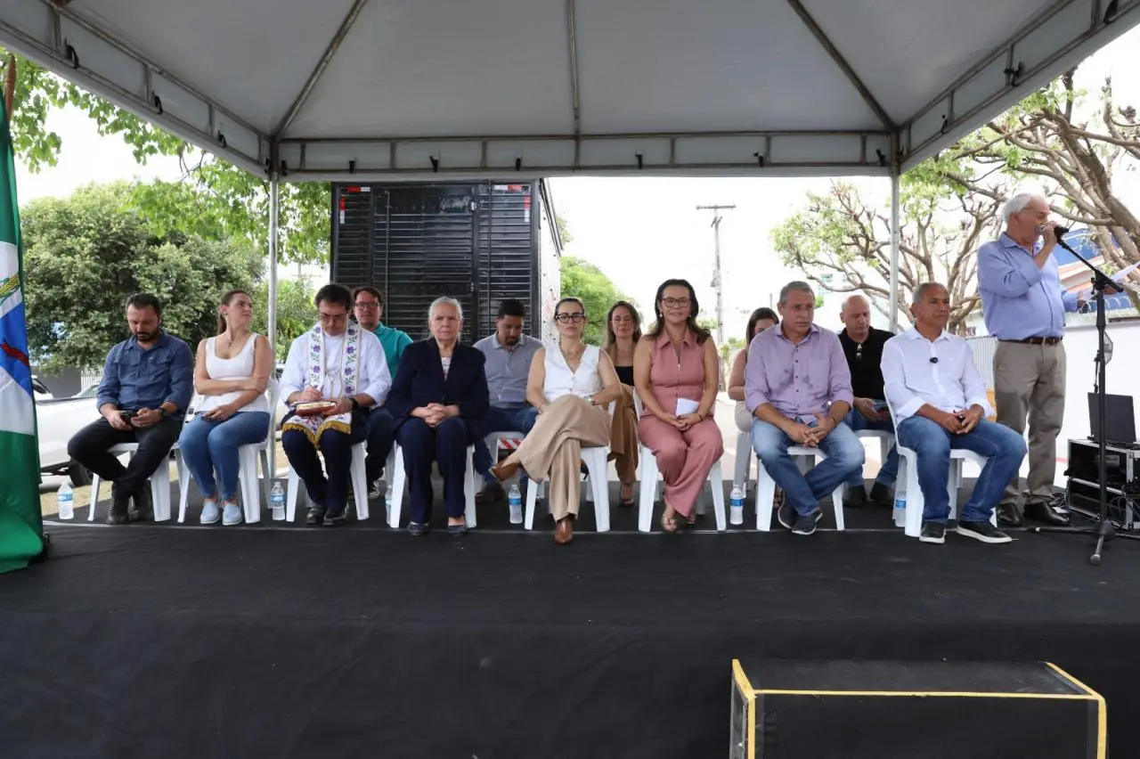 Autoridades e convidados estão sentados em cadeiras sobre um palco montado em área externa, coberto por tenda. Homens e mulheres de diferentes idades acompanham a solenidade, enquanto um homem fala ao microfone. O local tem árvores ao redor e clima institucional.