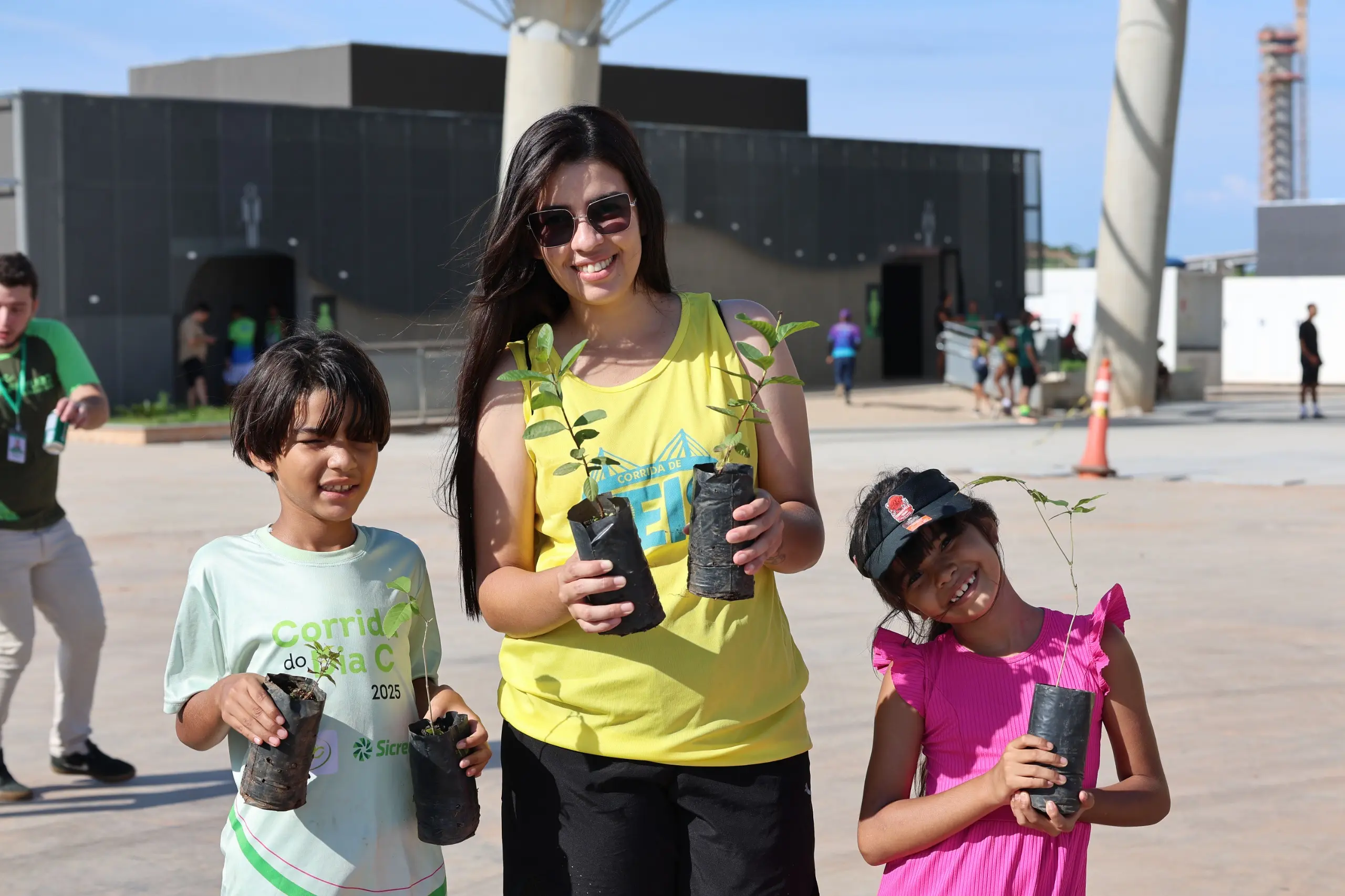Mulher e duas crianças posam em área externa do evento, sorrindo enquanto seguram mudas de árvores em sacos plásticos. Ao fundo, estrutura coberta, cones de sinalização e participantes circulam no espaço amplo e ensolarado.
