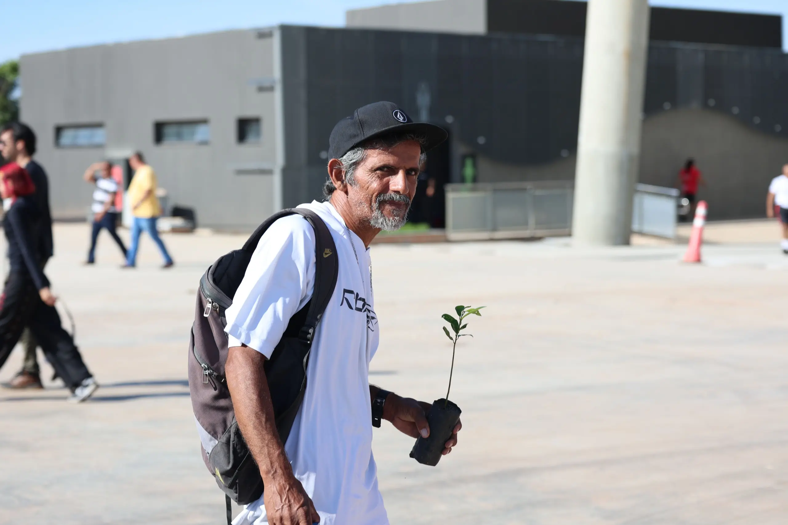 Homem com mochila caminha segurando uma muda de árvore. Ele atravessa o espaço do evento após a corrida, com outros participantes dispersos ao redor.
