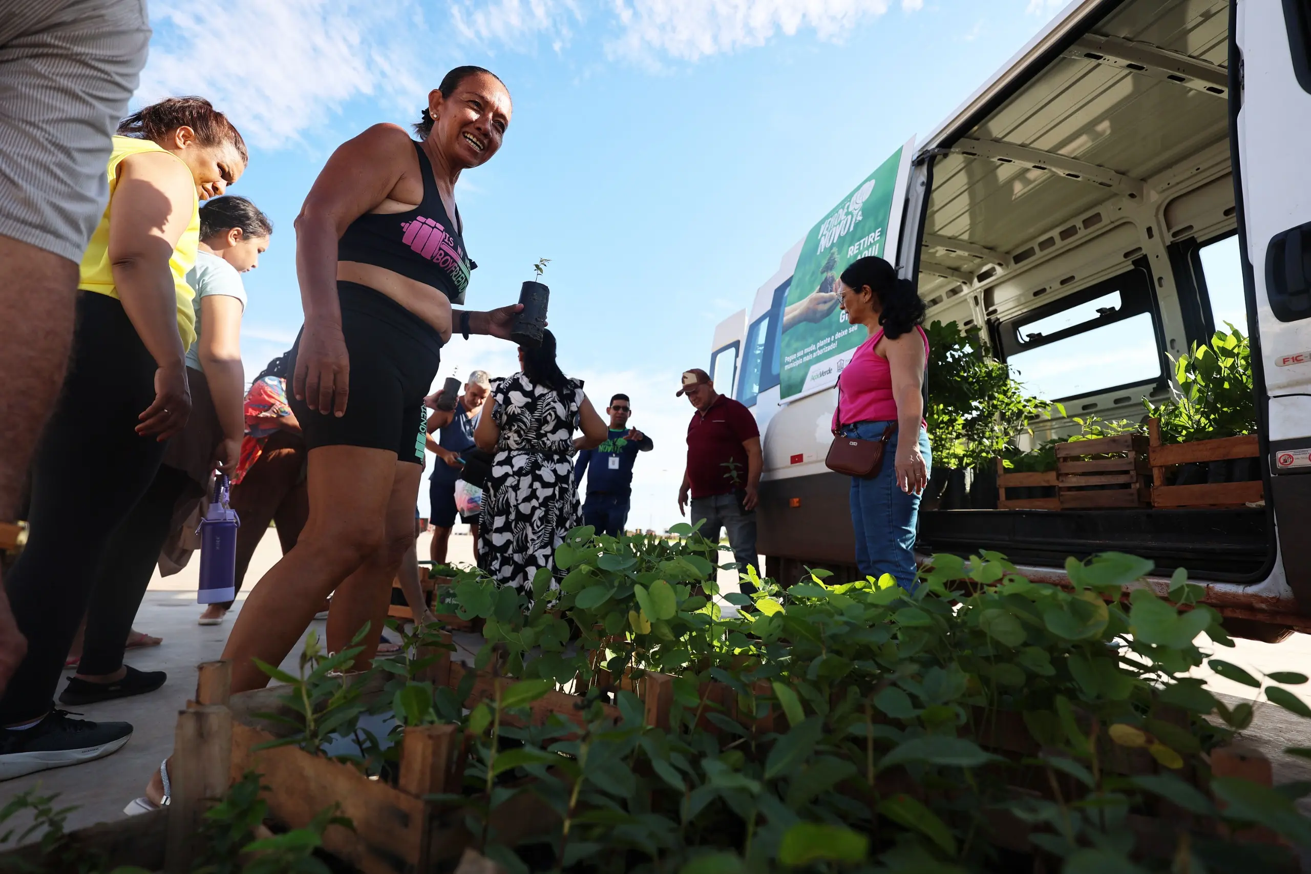 Participantes formam fila ao lado da van do Verde Novo. Pessoas com medalhas escolhem mudas dispostas em caixas, sob sol forte, com estrutura do complexo ao fundo.