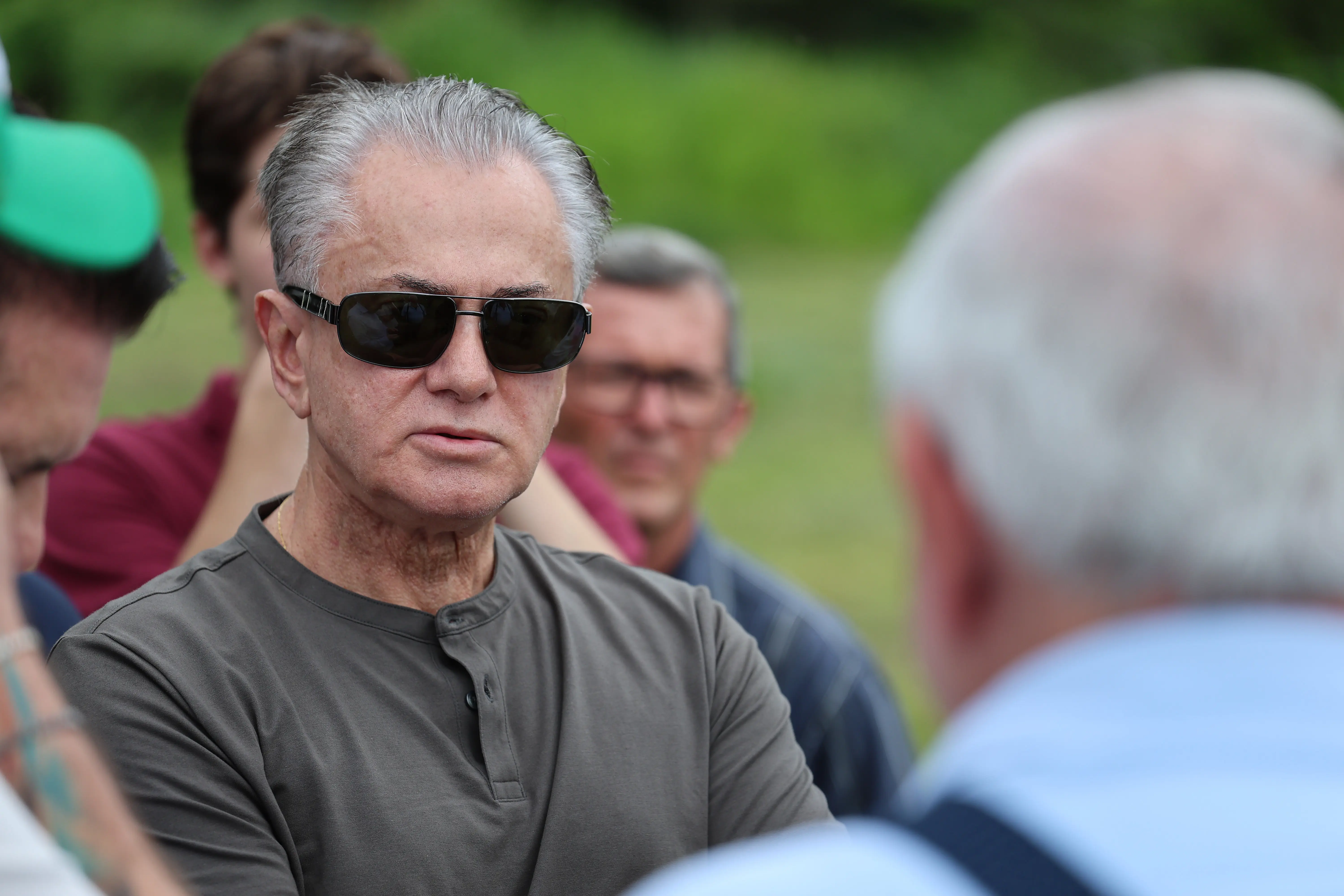 Close-up de um senhor, cabelos grisalhos, usando óculos de sol escuros e uma camisa cinza escura de gola henley. Ele está ao ar livre, em uma conversa com outras pessoas.