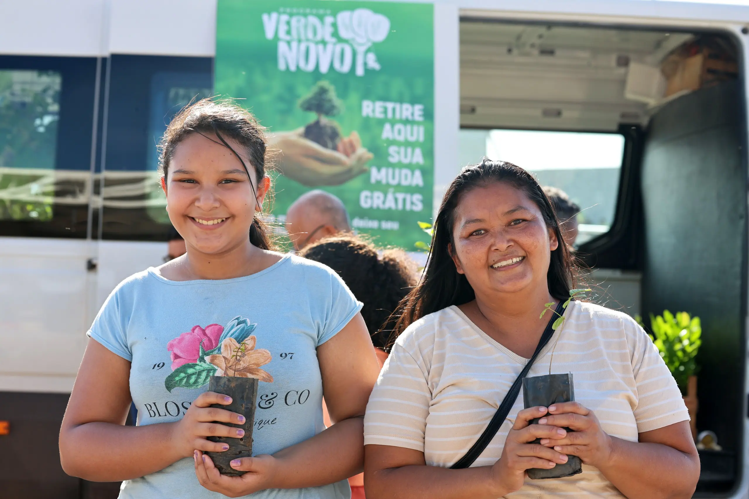 Mulher e criança sorriem para a câmera enquanto seguram mudas de árvores. A foto é registrada em área aberta do evento, com circulação de atletas ao fundo.