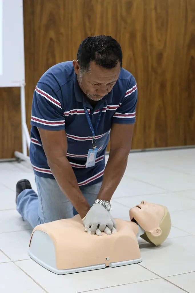 Foto vertical do João Carlos Machado durante um treinamento de reanimação cardiopulmonar, feita em um boneco, na sala de aula da Escola da Magistratura. Ele é um homem negro, de cabelo preto, barba grisalha, usando calça jeans, camiseta polo azul com listras brancas e vermelhas e crachá funcional. 