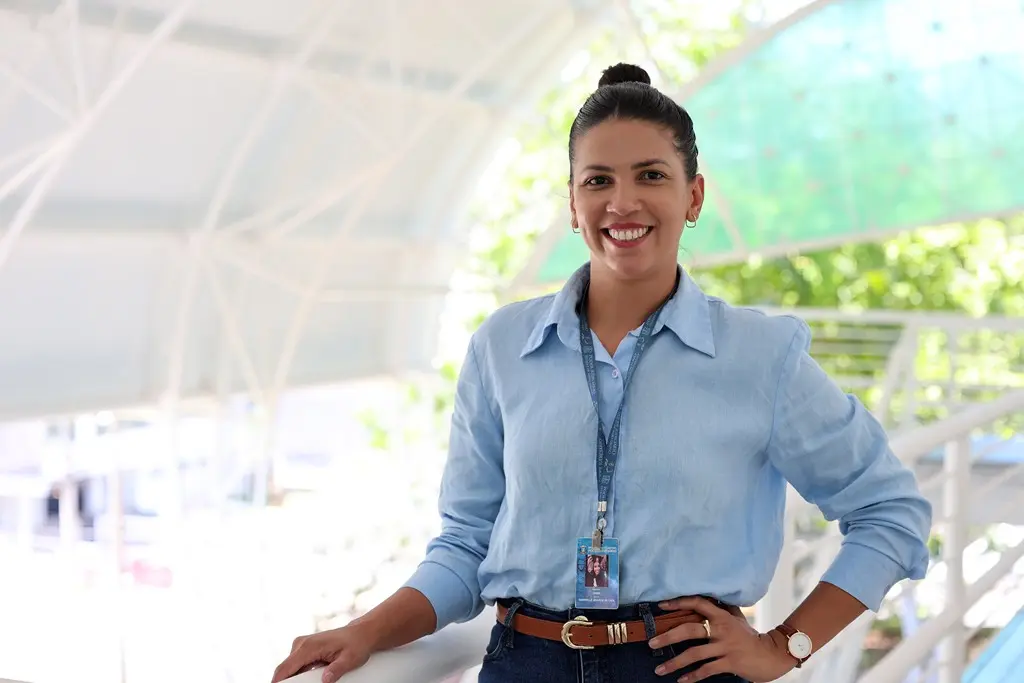 Foto horizontal da Gabrielle Soares posando sorrindo, com a mão apoiada no corrimão. Ela é uma jovem alta de pele parda, cabelos e olhos castanhos escuros, cabelos estão presos em coque. Ela usa calça jeans, camisa de manga comprida azul clara e crachá. 
