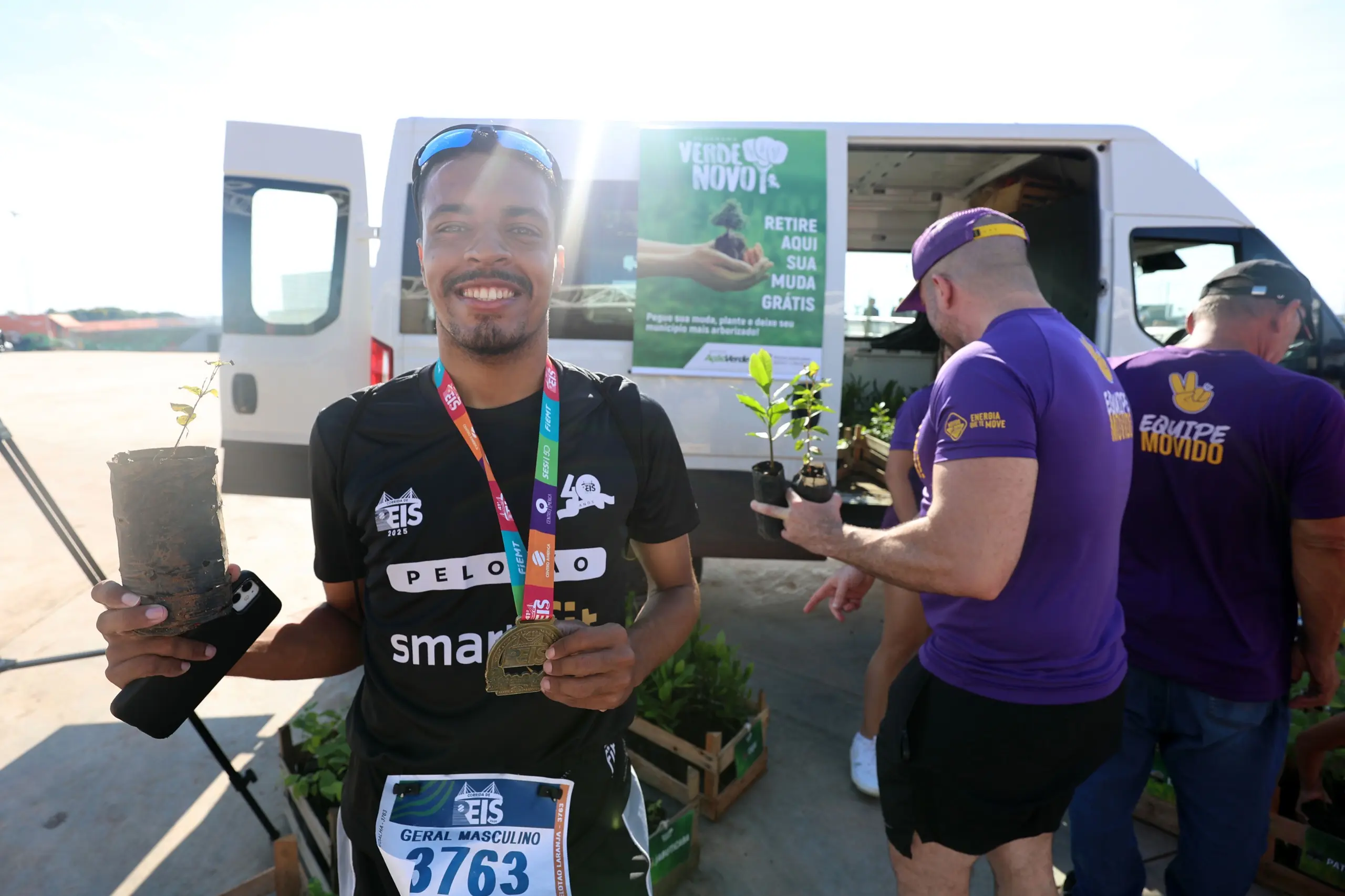 Homem de camiseta preta e medalha posa segurando uma muda. Ao fundo, pessoas caminham pelo local após a prova, com a estrutura coberta visível.