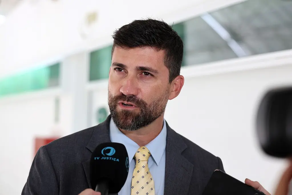 Foto horizontal do juiz Vinícius Paiva Galhardo durante entrevista à TV Justiça. Ele é um homem branco, de olhos, cabelos e barba castanhos, usando camisa azul clara, gravata amarela e terno cinza. 
