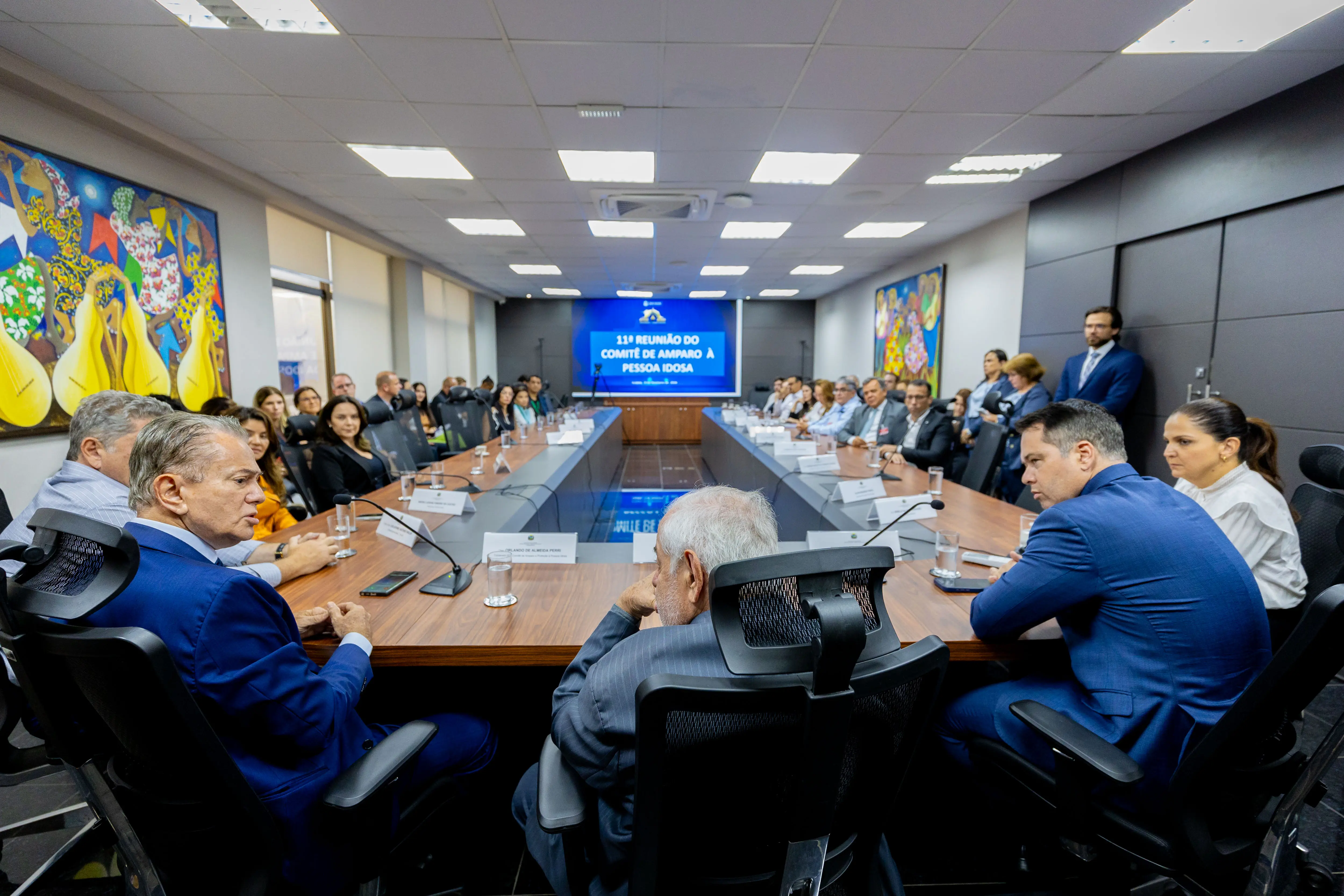 Reunião em sala de conferência com várias pessoas sentadas em volta de mesa em U. Tela ao fundo exibe 