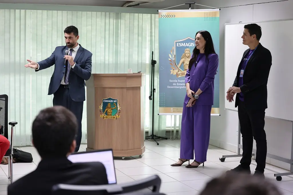 Foto horizontal em plano aberto que mostra a sala de aula da Escola da Magistratura com juízes sentados e. À frente, em pé, estão: o juiz Vinicius Paiva falando ao microfone, a juíza Joseane Antunes e o servidor Thomas Caetano.