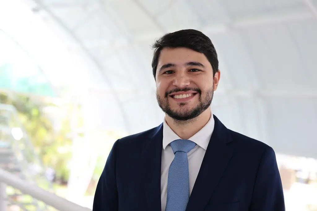 Foto horizontal em plano fechado do juiz Victor Hugo Sousa Santos sorrindo para a foto, no pátio da Escola da Magistratura. ele é um homem jovem, branco, de cabelos, barba e olhos castanhos, usando camisa branca, gravata azul clara e terno azul marinho.