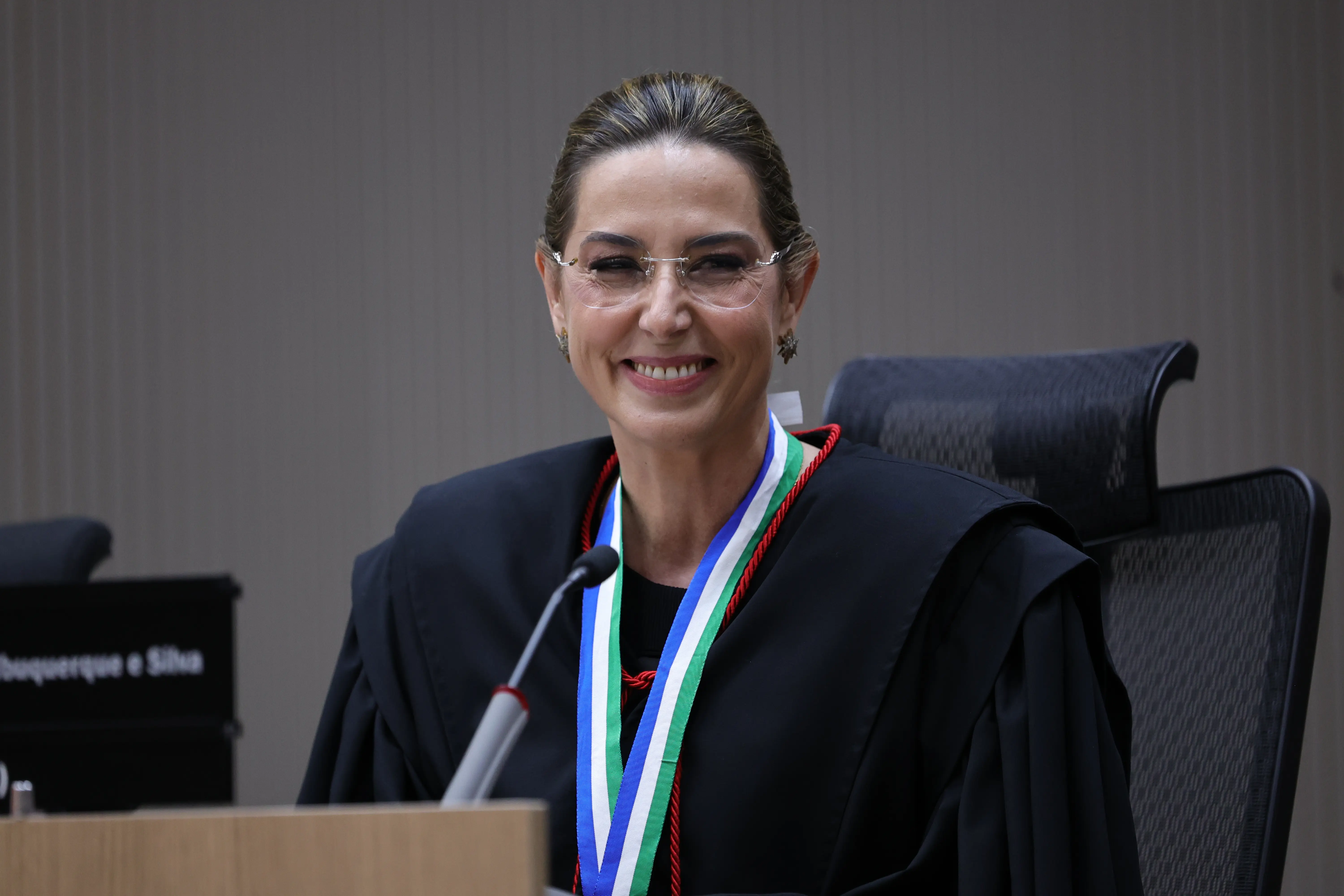 Mulher sorri sentada à bancada usando traje de magistrado e medalha nas cores verde, branco e azul no pescoço. Ao lado, microfone sobre a mesa. Ambiente de tribunal com parede cinza ao fundo.