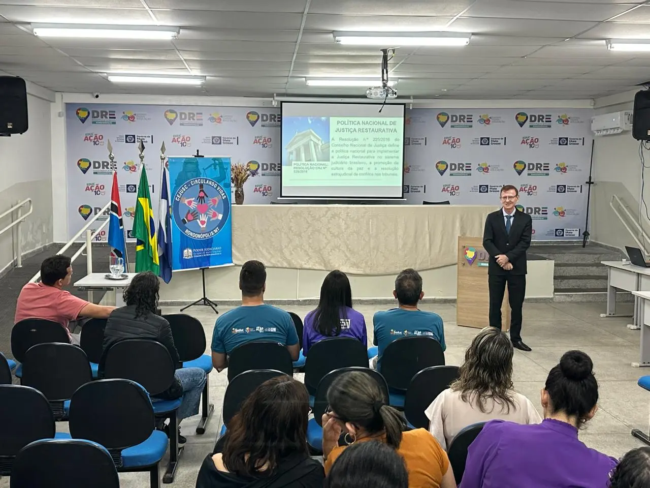 A foto mostra um auditório com pessoas sentadas em cadeiras azuis, assistindo a uma palestra sobre Justiça Restaurativa. Um juiz está à frente, ao lado de bandeiras e de um telão com apresentação institucional.