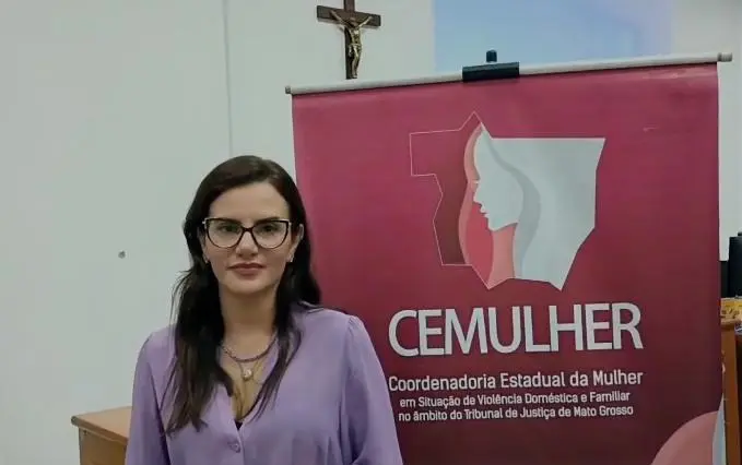 Uma mulher de óculos, cabelos escuros e blusa lilás posa em frente ao banner do CEMULHER/TJMT.