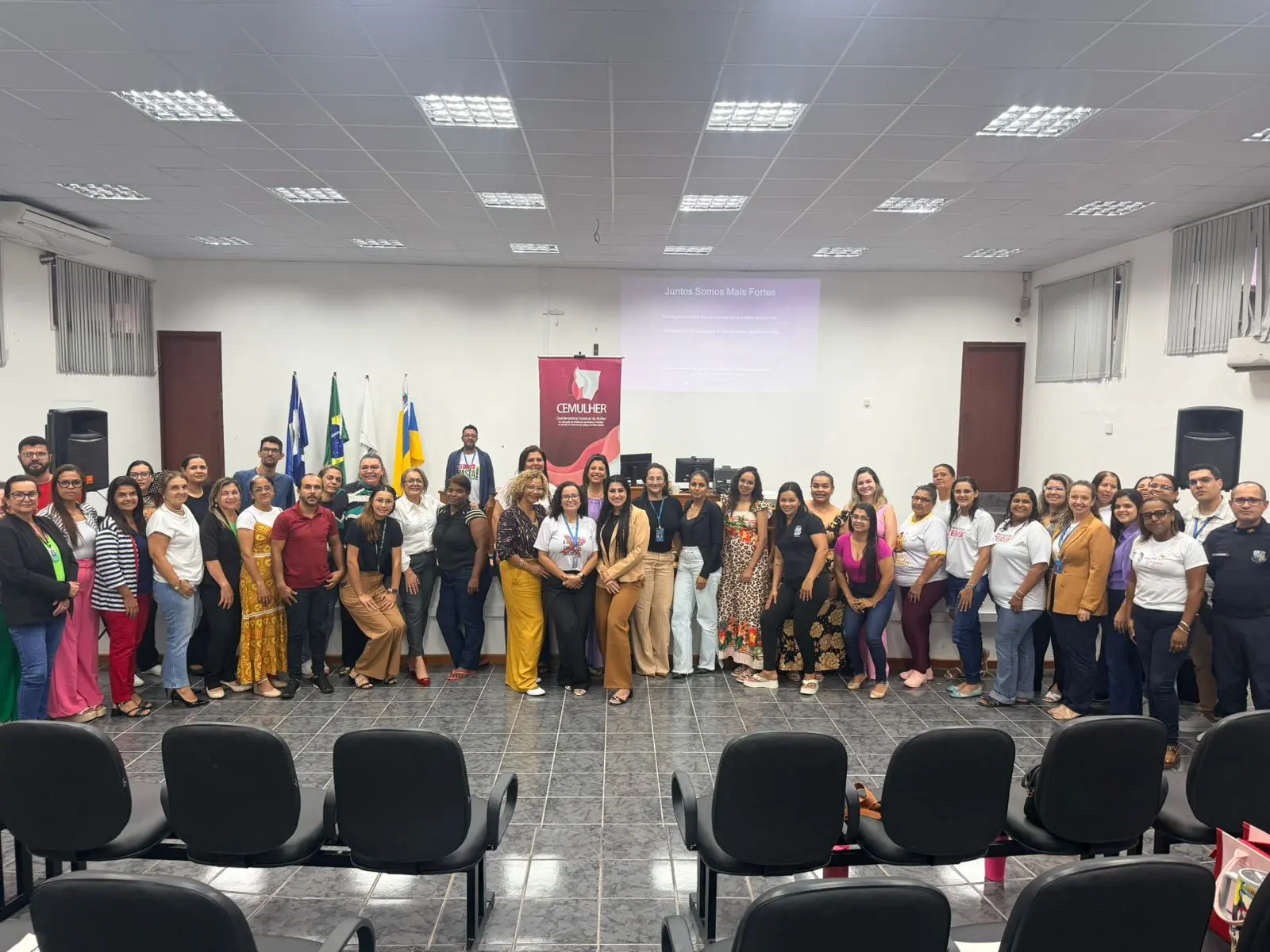 Grupo de cerca de 40 pessoas posadas em salão com bandeiras e banner do CEMULHER ao fundo. Na tela, a frase 