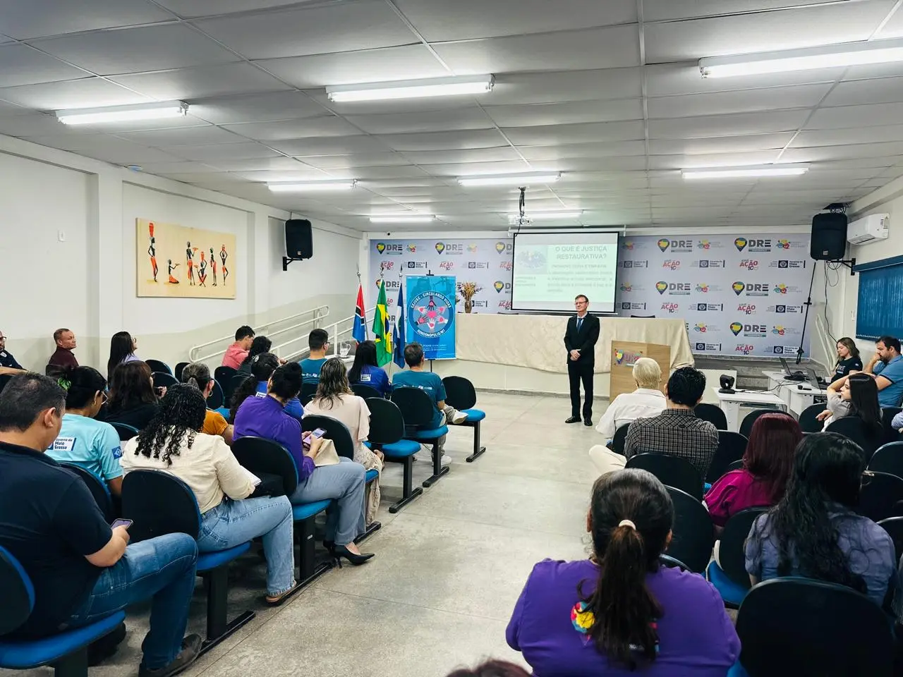 A foto apresenta uma visão ampla do auditório lotado, com público atento à palestra. O palestrante está em pé próximo ao púlpito, enquanto o telão exibe o tema do encontro, em um espaço organizado e iluminado.