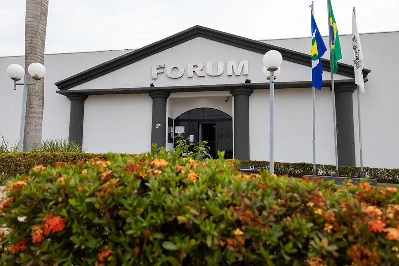 Foto horizontal em plano aberto que mostra a fachada do Fórum de Peixoto de Azevedo. é um prédio em estilo que remete às fachadas greco-romanas, com mastros com as bandeiras do Brasil, de Mato Grosso e do Poder Judiciário e flores na frente do prédio.