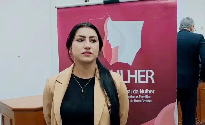 Uma mulher de cabelos pretos longos, veste blazer bege sobre blusa preta. Ao fundo, banner vinho do CEMULHER/TJMT.