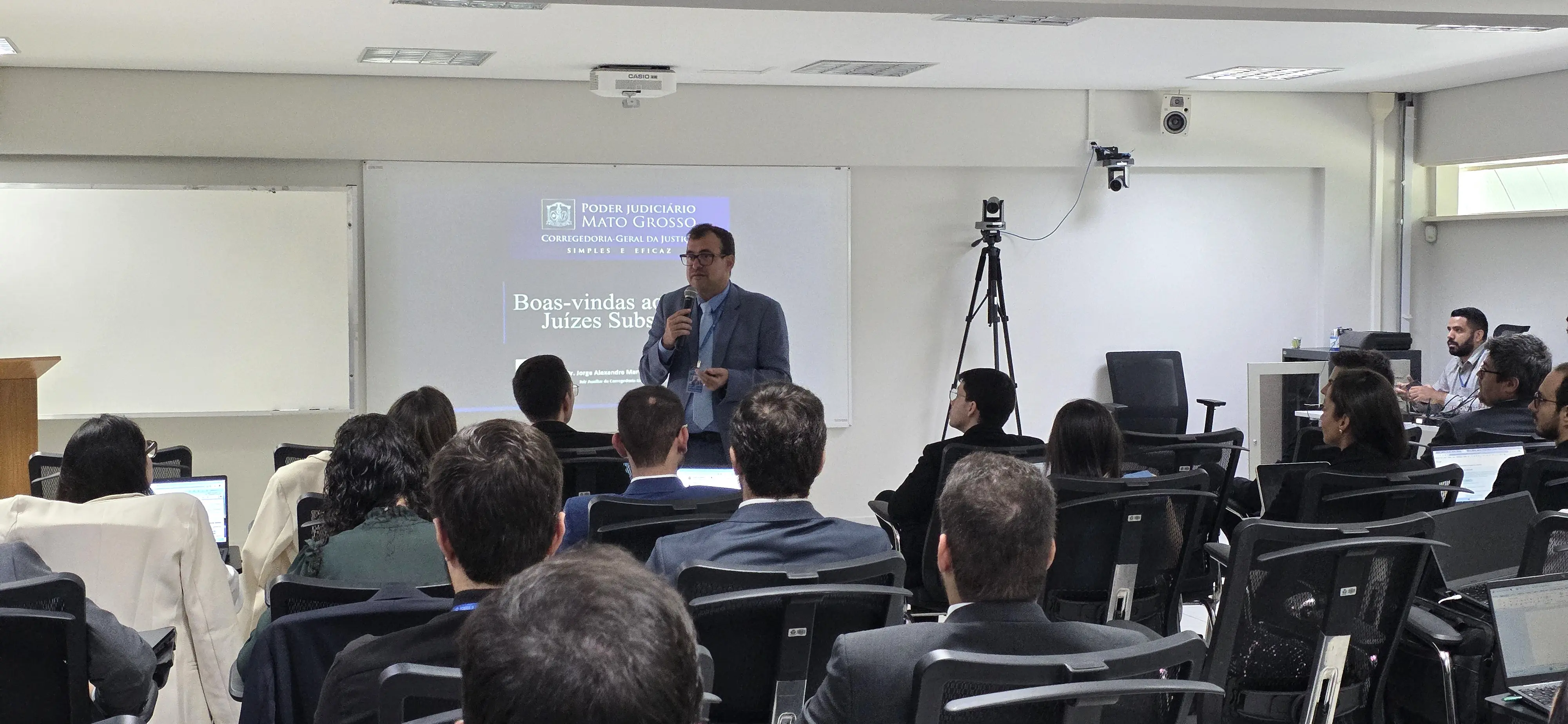 Foto do juiz auxiliar da Corregedoria, Jorge Alexandre Martins Ferreira. Homem de pele clara, cabelo curto escuro e óculos, veste terno azul, camisa clara e gravata. Está em pé, segurando um microfone, enquanto fala aos magistrados sentados em cadeiras pretas 