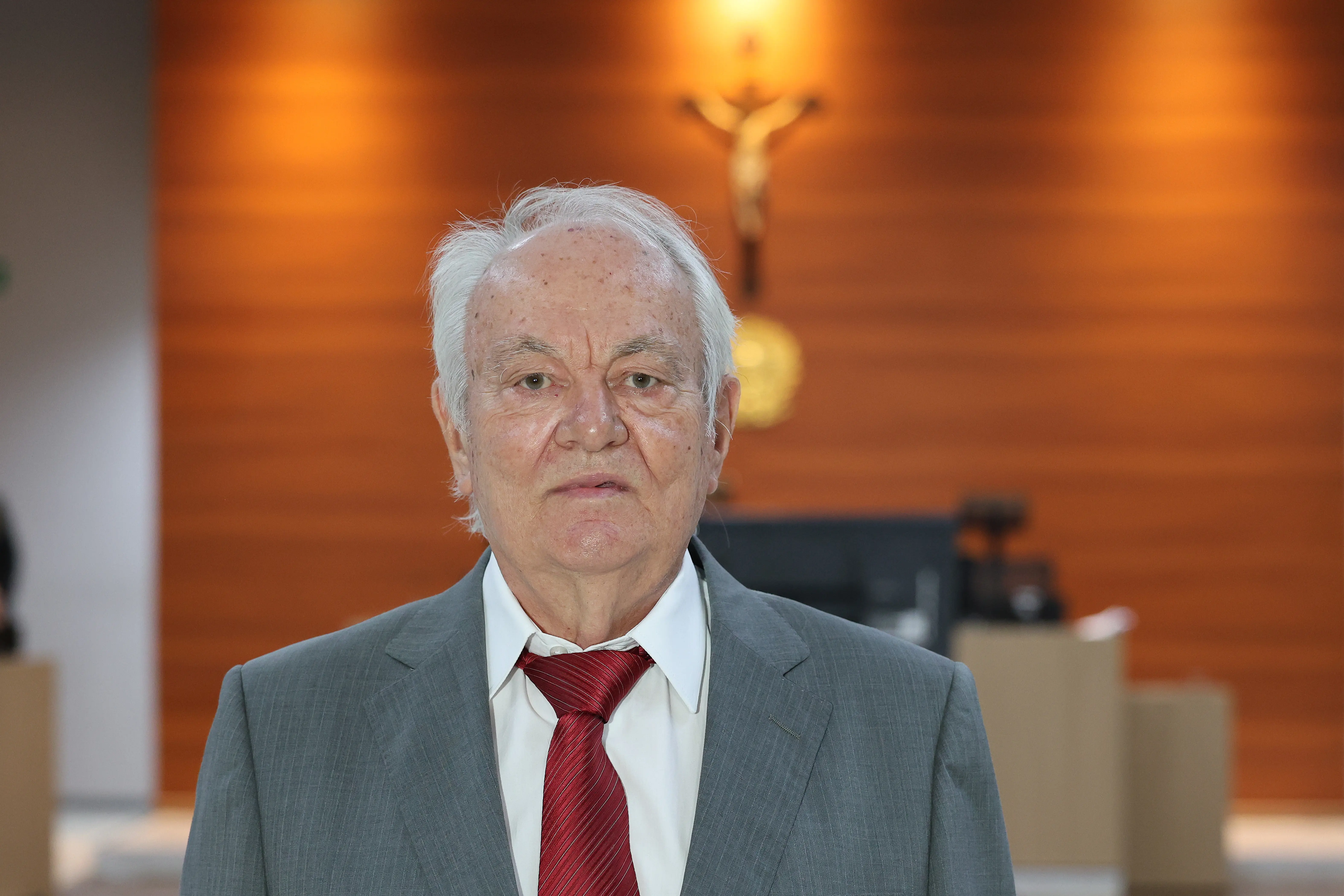 Um homem idoso de pele clara e cabelos brancos, vestindo terno cinza, camisa branca e gravata vermelha, posa para a foto. Ao fundo, uma parede de madeira com um crucifixo e o brasão nacional.