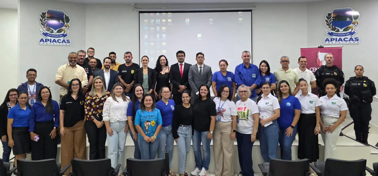 Foto em grupo da Rede de Enfrentamento em Apiacás. Homens e mulheres posam à frente de um telão, ladeados pelo brasão da prefeitura e o banner da CEMULHER