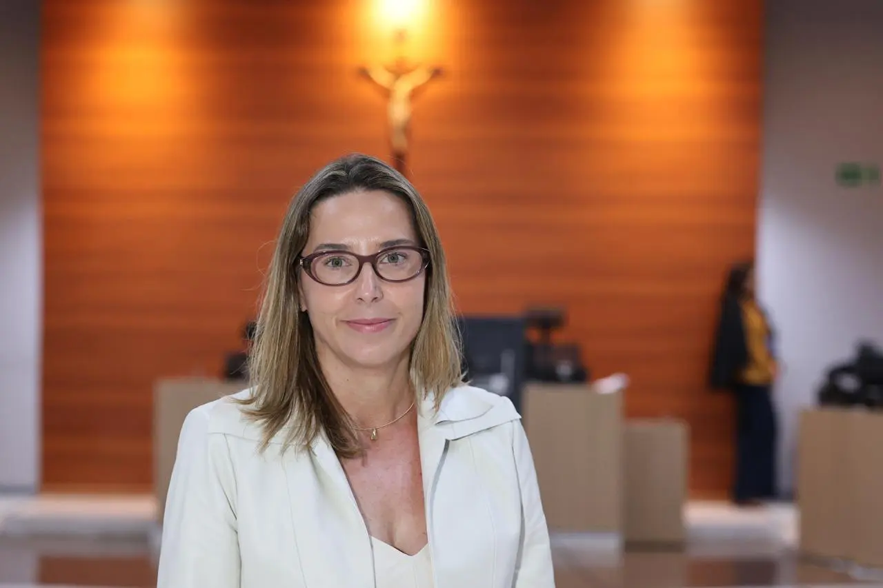 Uma mulher de pele clara e óculos, vestindo blazer branco, posa para a foto. Ao fundo, uma parede de madeira com um crucifixo e o brasão nacional em dourado em um ambiente jurídico.
