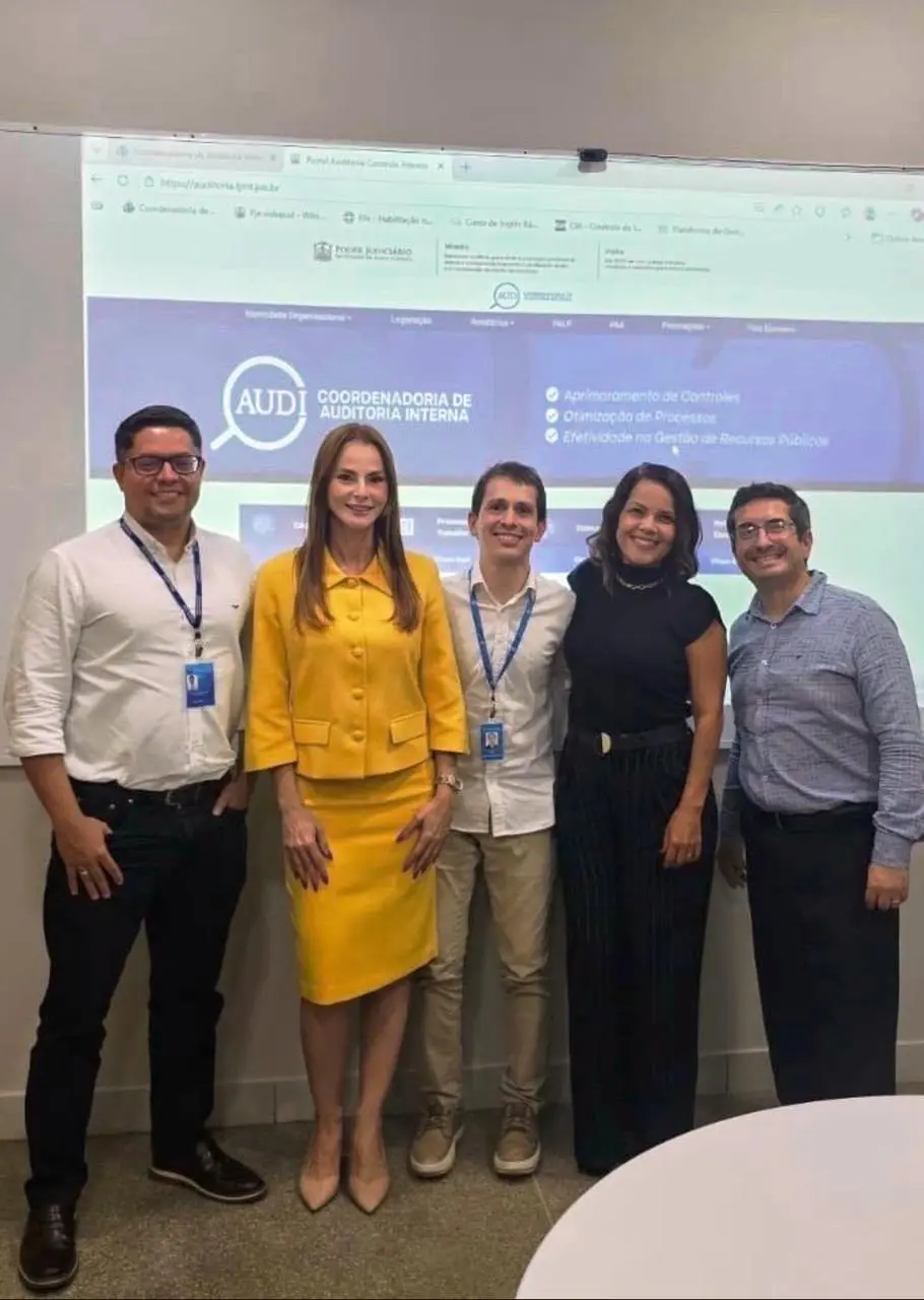 Cinco profissionais posam diante de tela com site da Coordenadoria de Auditoria Interna. Em trajes formais, exibem crachás e postura institucional, em registro coletivo no ambiente de trabalho.