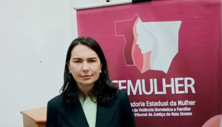 Uma mulher de cabelos escuros e lisos, veste blazer preto sobre blusa verde-clara. Ao fundo, um banner vinho com o logotipo do CEMULHER/TJMT.