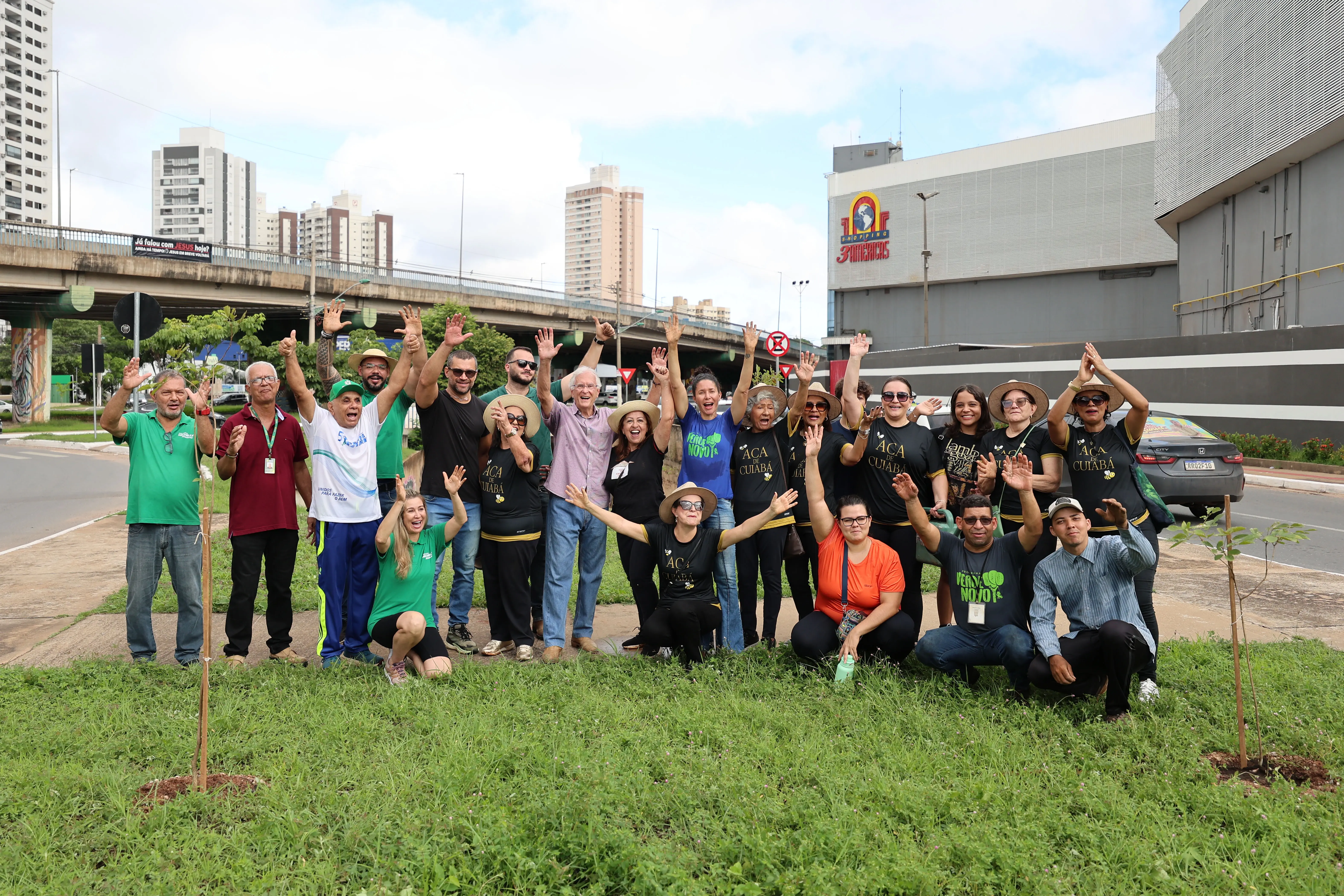 Grupo de cerca de 20 pessoas sorri e ergue os braços em área gramada de avenida urbana, com viaduto e prédios ao fundo. Mudas recém-plantadas aparecem no chão.