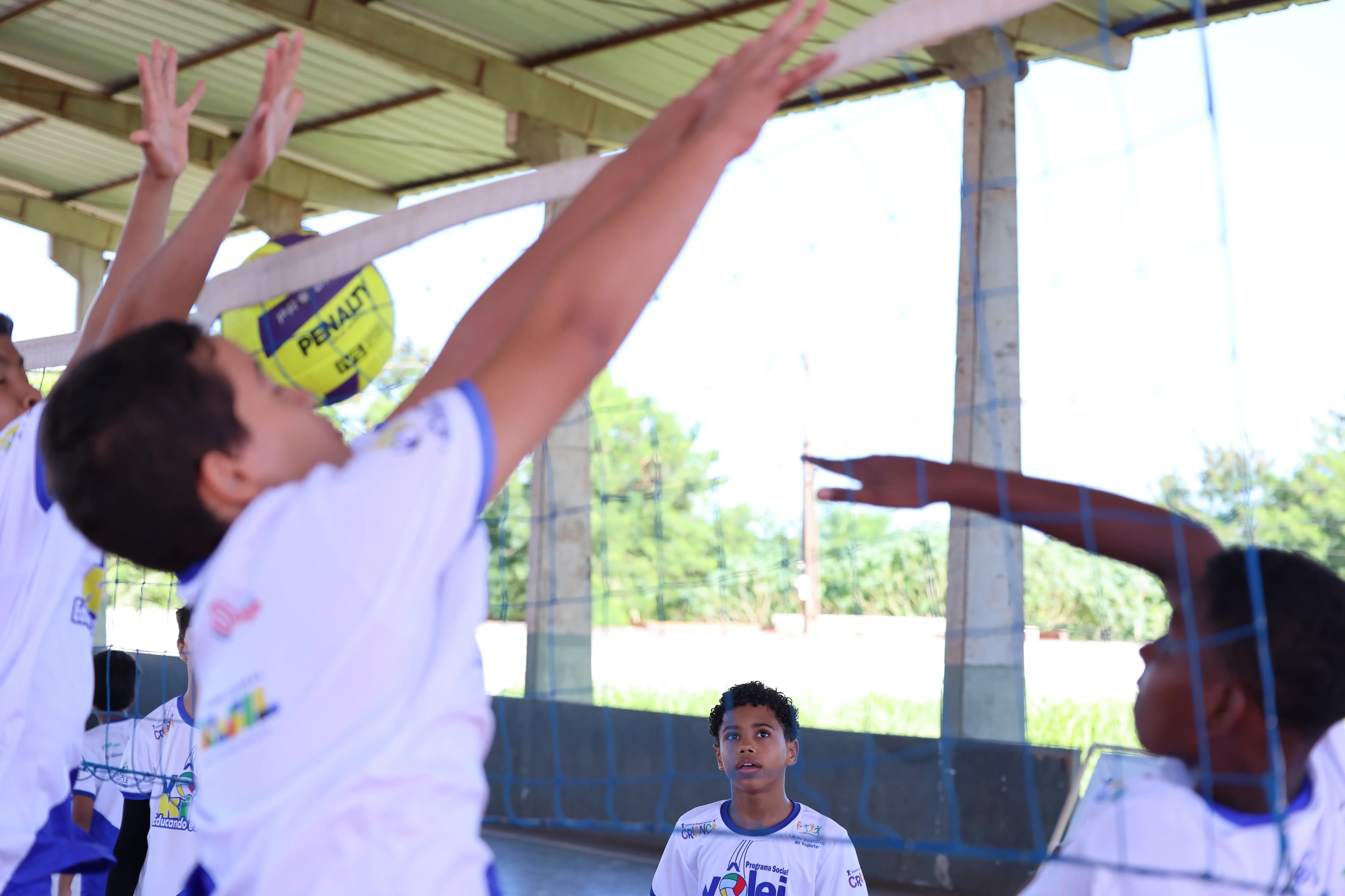 Criança salta para arremessar bola em atividade esportiva. Outros colegas aparecem ao redor observando.