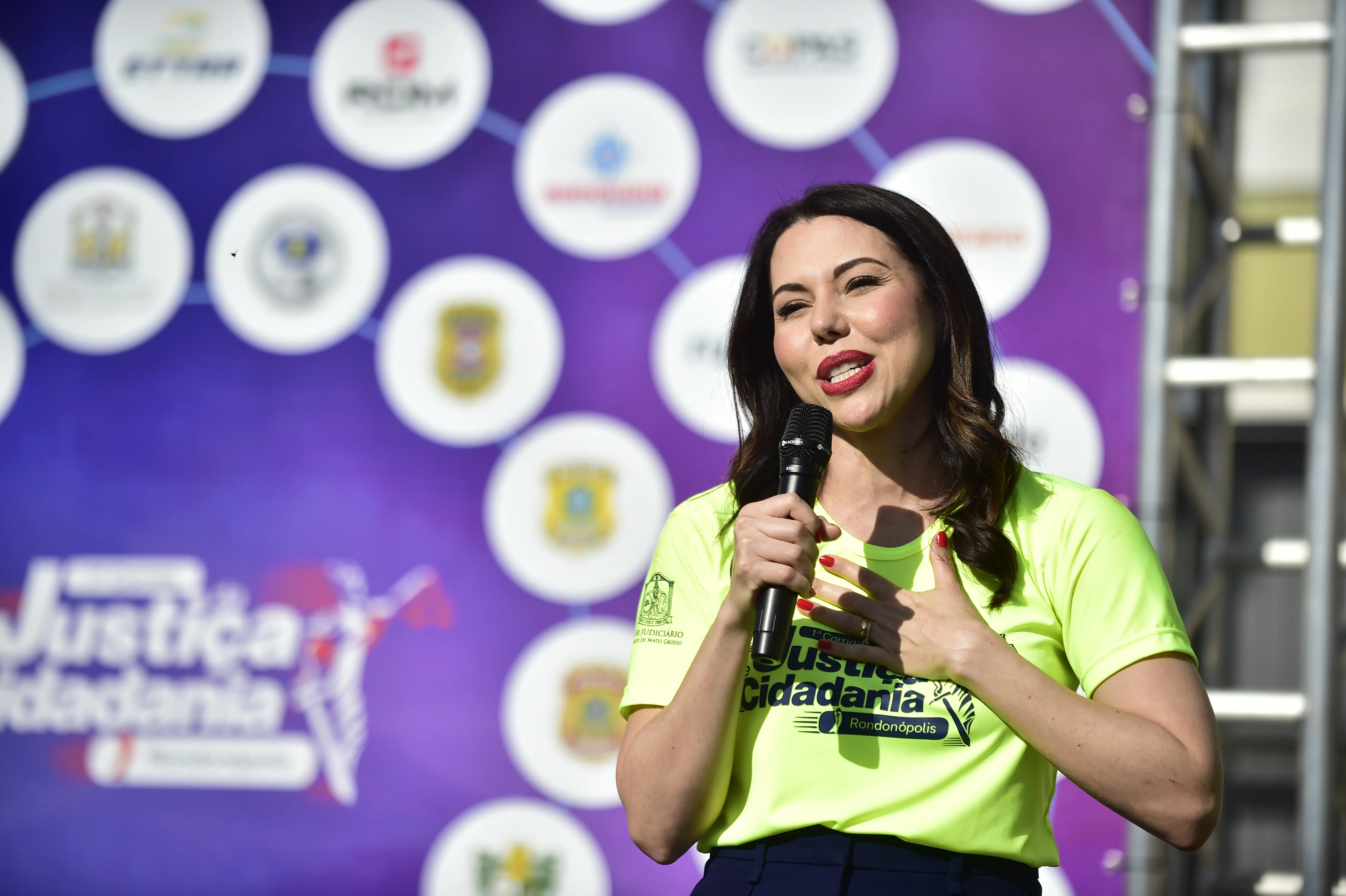 Uma mulher em destaque, vestindo camiseta verde limão do evento, fala ao microfone emocionada em frente a um painel de logos.
