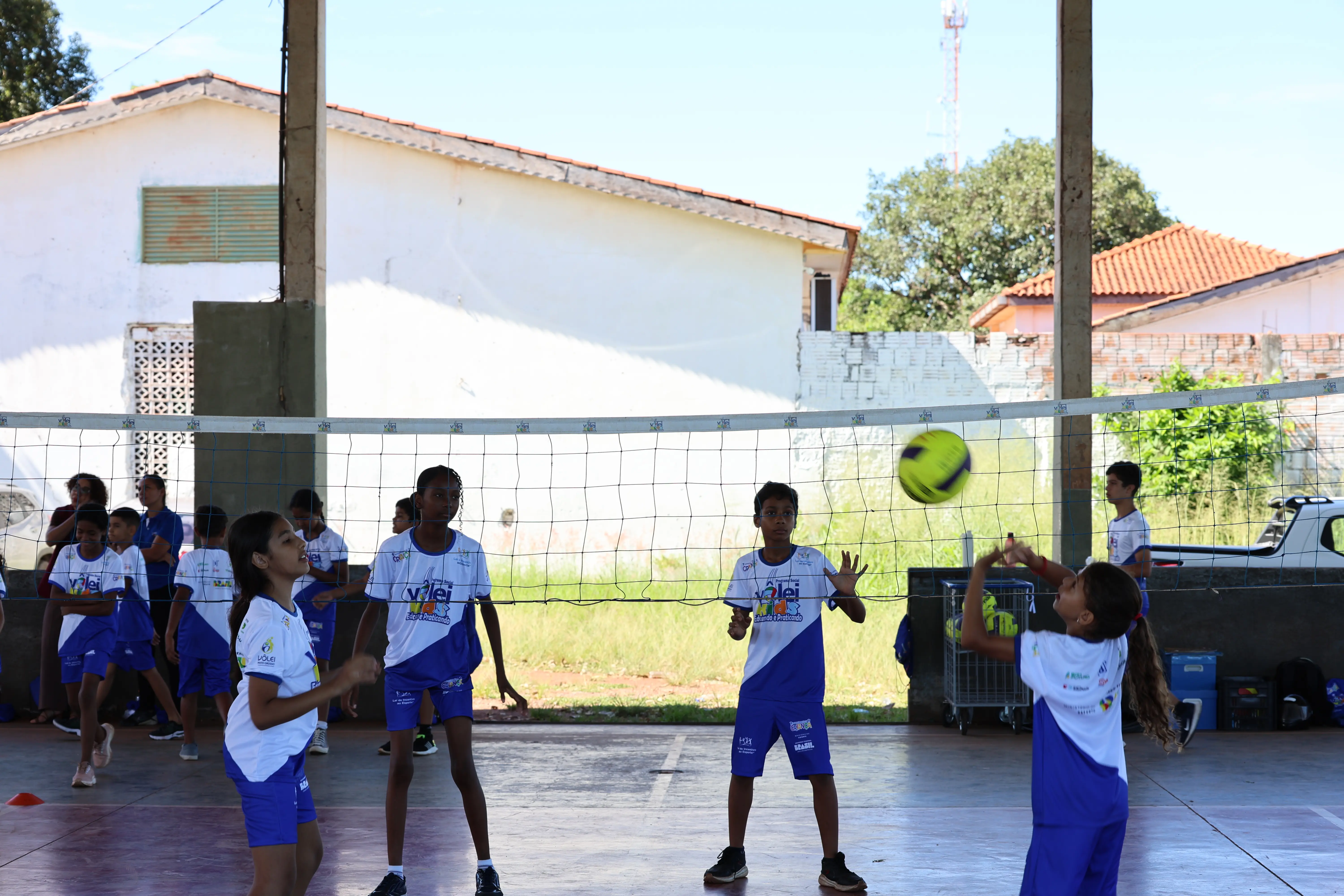 Grupo maior de alunos em quadra externa coberta. Crianças espalhadas, algumas conversam, outras brincam.
