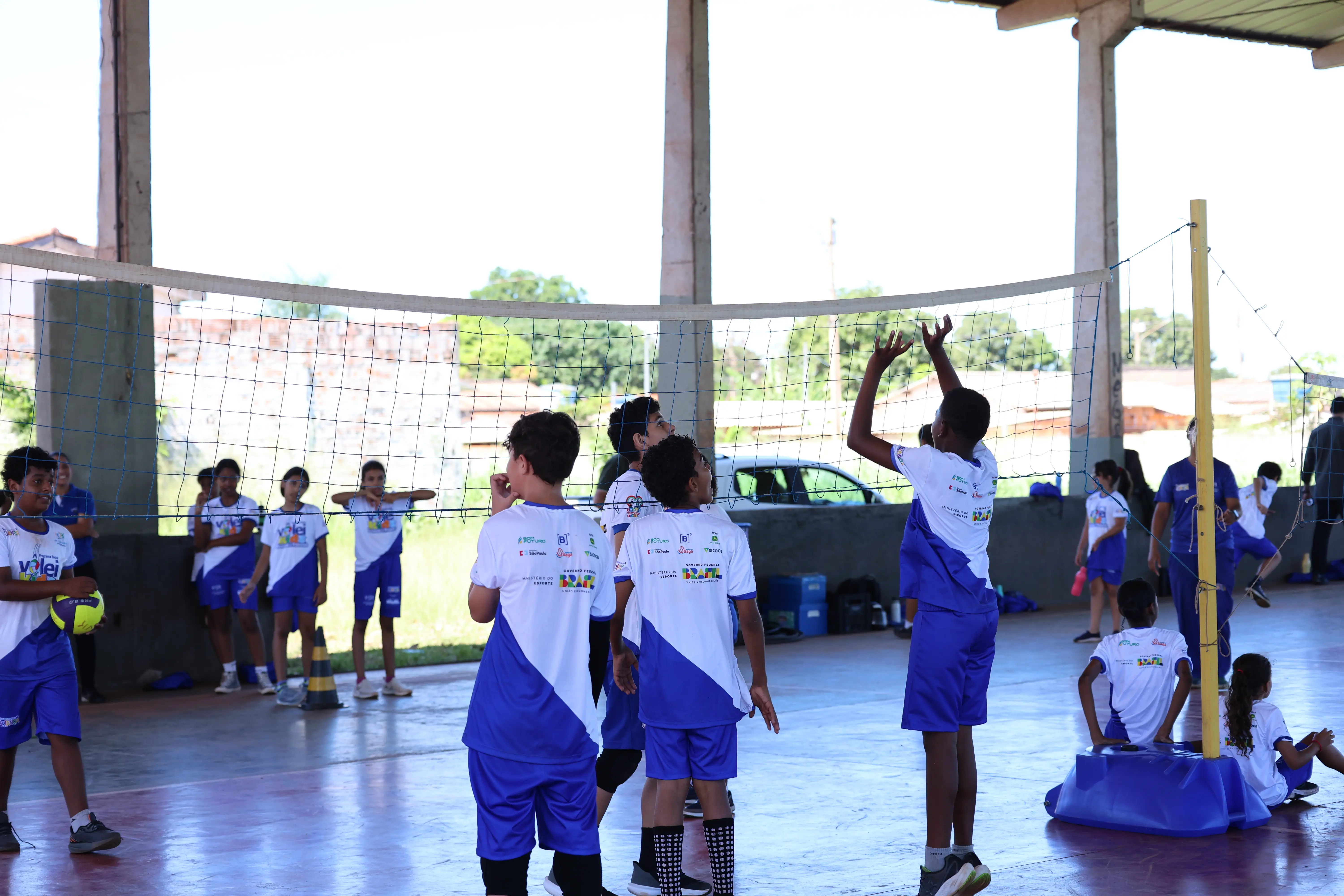 Crianças reunidas próximas à lateral da quadra, algumas em pé e outras em movimento, aguardando ou participando da atividade.