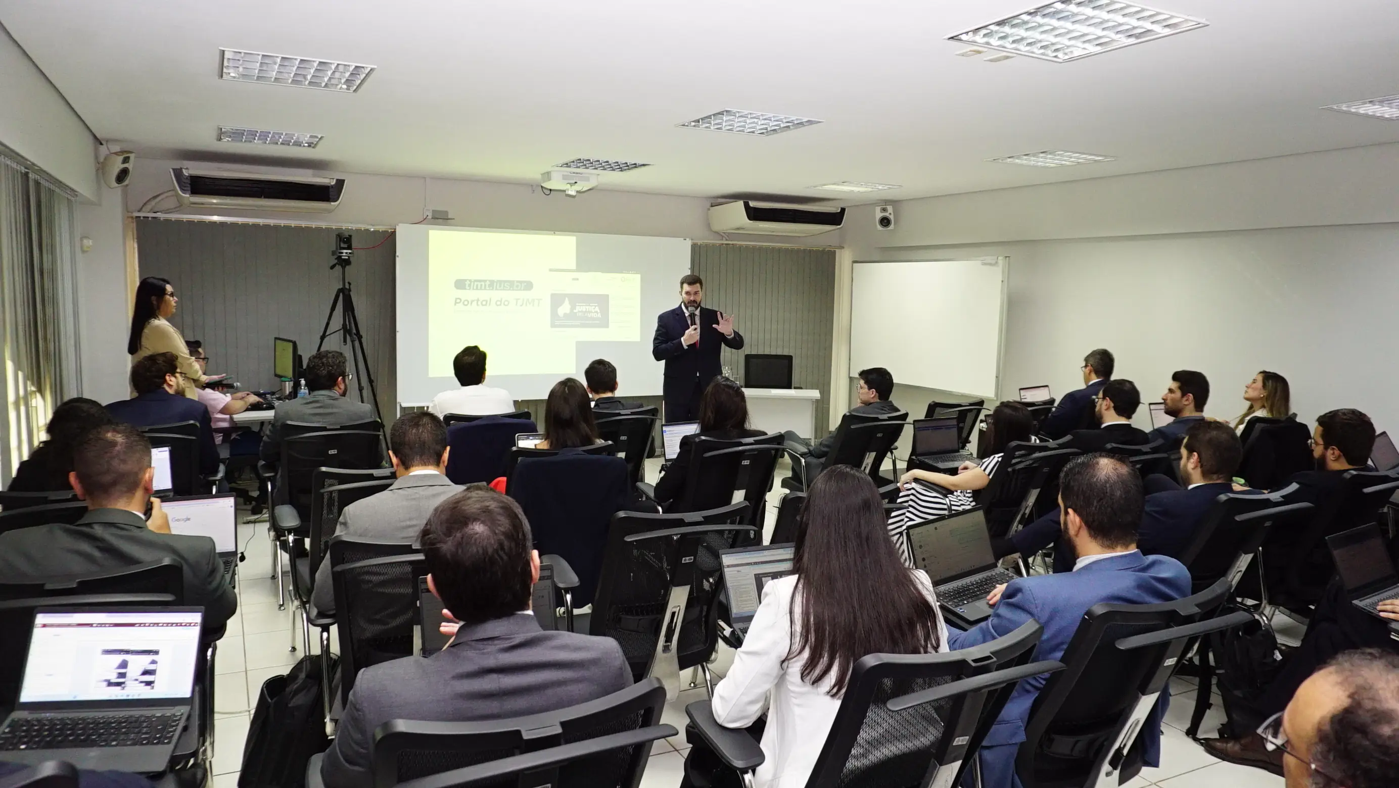Ccom no Cofi: Vista ampla de uma sala de treinamento com público sentado em cadeiras pretas, muitos utilizando notebooks. À frente, um homem de terno preto fala ao microfone ao lado de um grande telão.