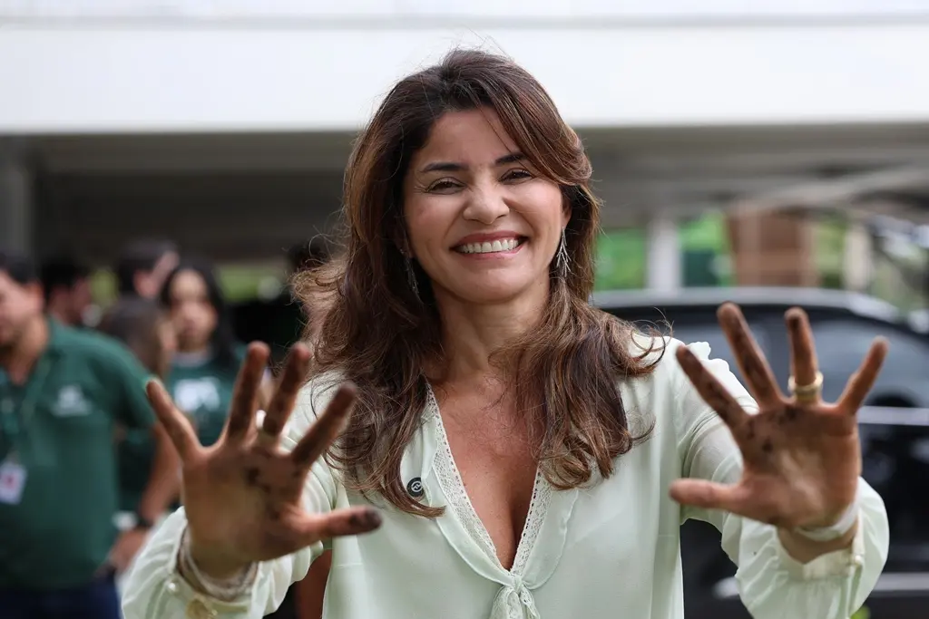 Foto horizontal colorida, em plano fechado, que mostra a defensora pública-geral do Estado, Luziane Castro, posando para a foto sorrindo e mostrando as mãos sujas de terra, após plantar muda de árvore ao lado da sede da Defensoria. Ela é uma mulher parda, de longos cabelos castanhos e ondulados.