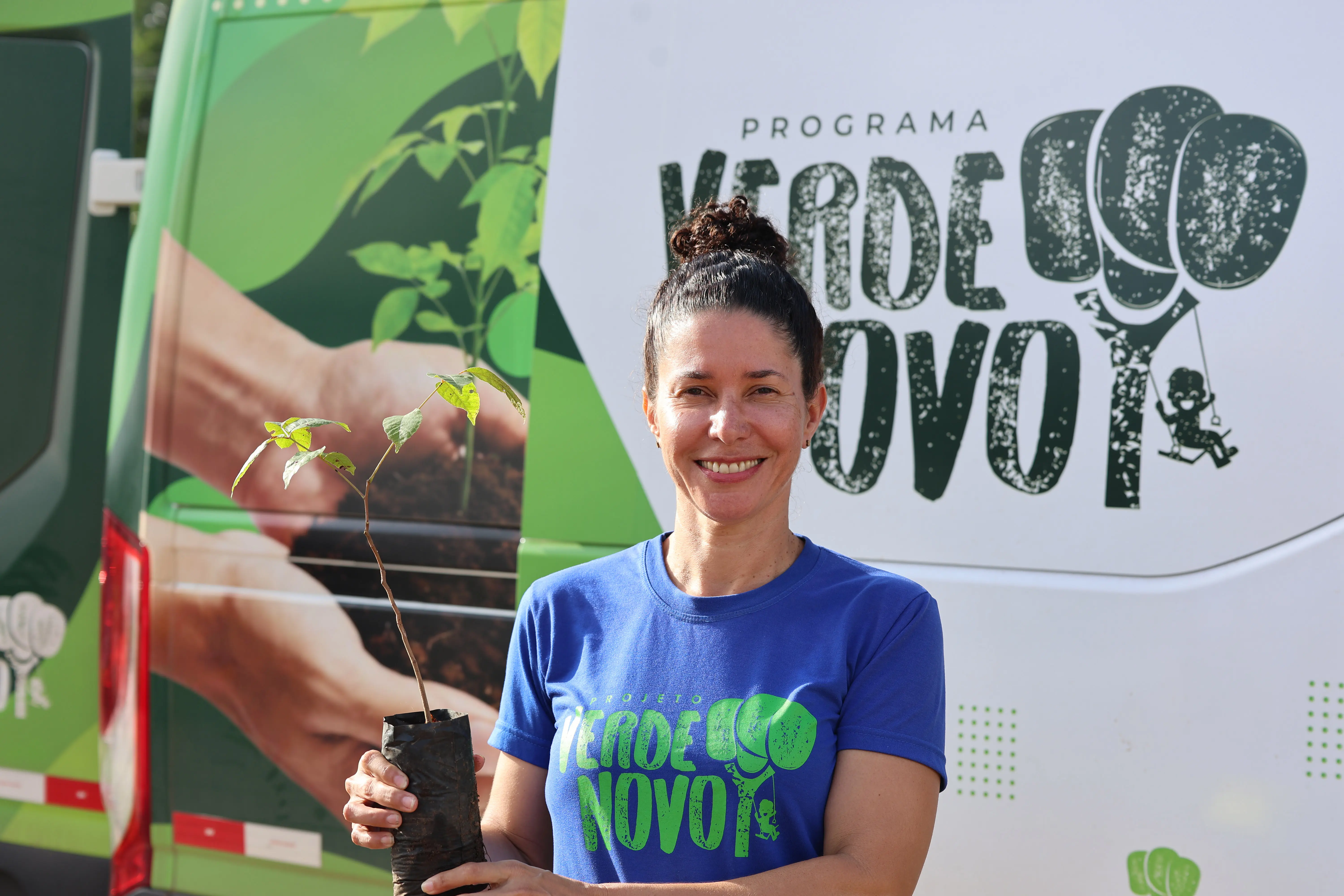Mulher de cabelos presos, camiseta azul com logotipo do Programa Verde Novo, segura uma muda. Ao fundo, van do programa estacionada.