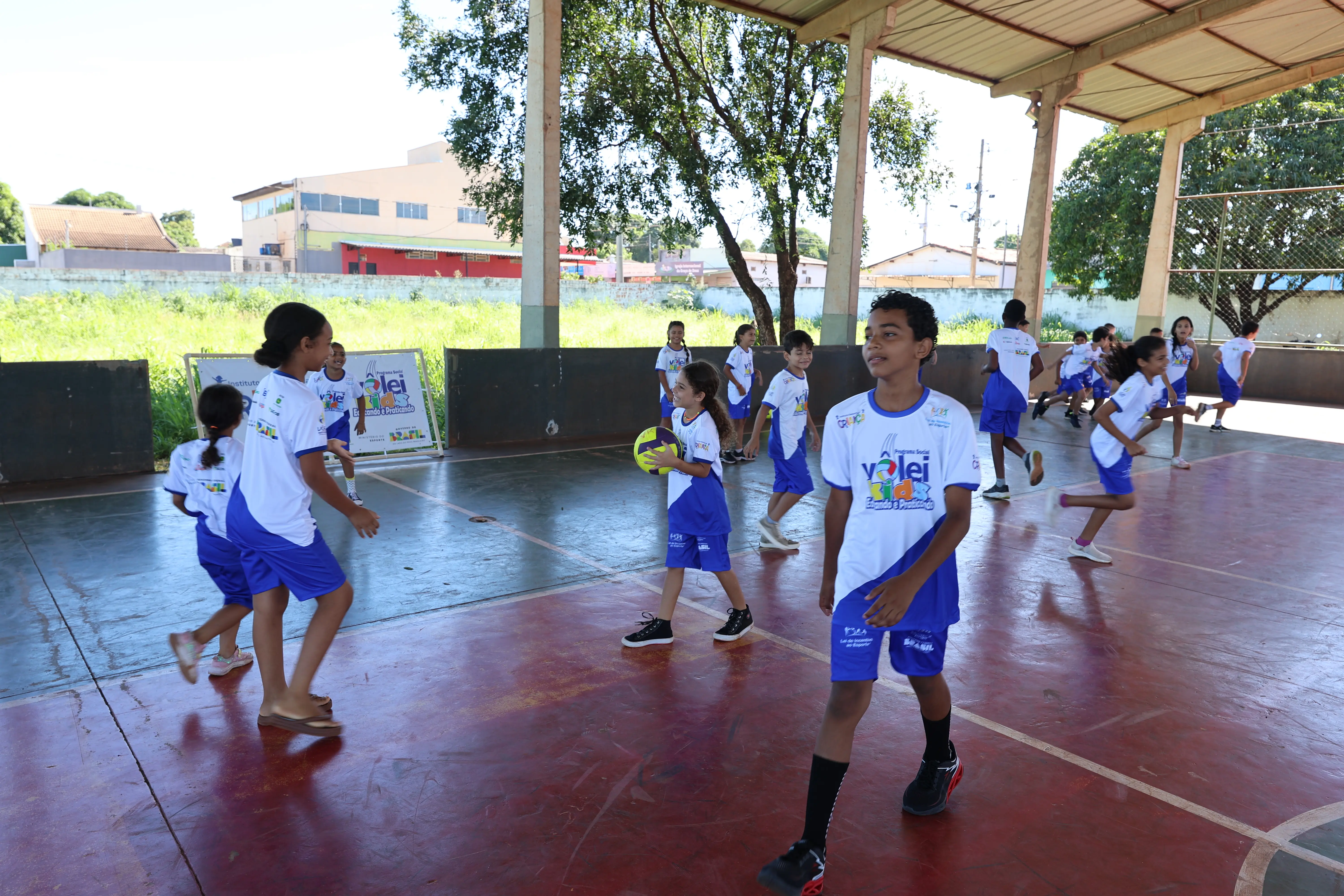 Crianças correm e brincam em quadra coberta. Movimento e interação marcam a cena, com uniforme esportivo.