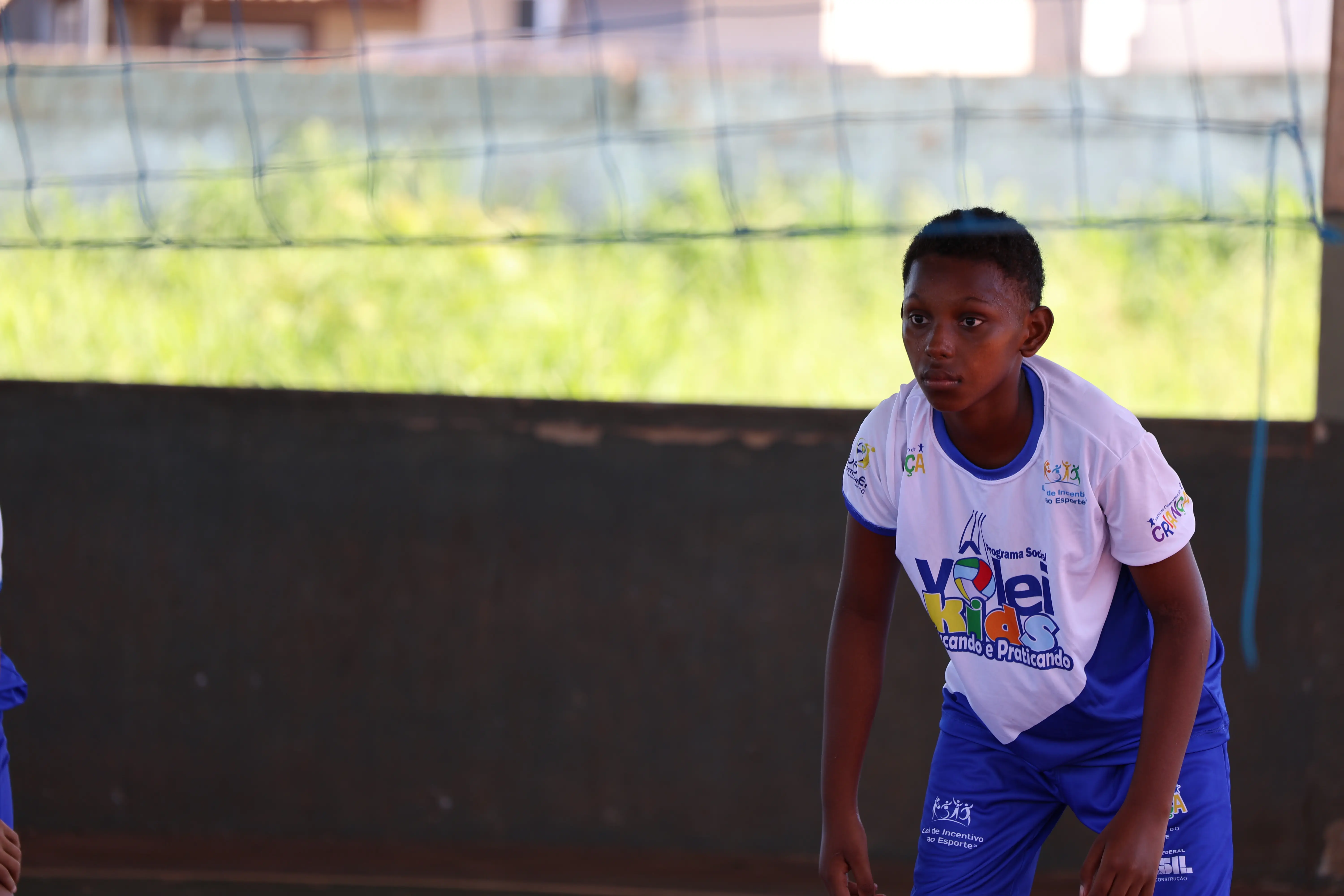 Menino em destaque segura bola, com uniforme escolar. Fundo desfocado de quadra, foco na expressão concentrada.