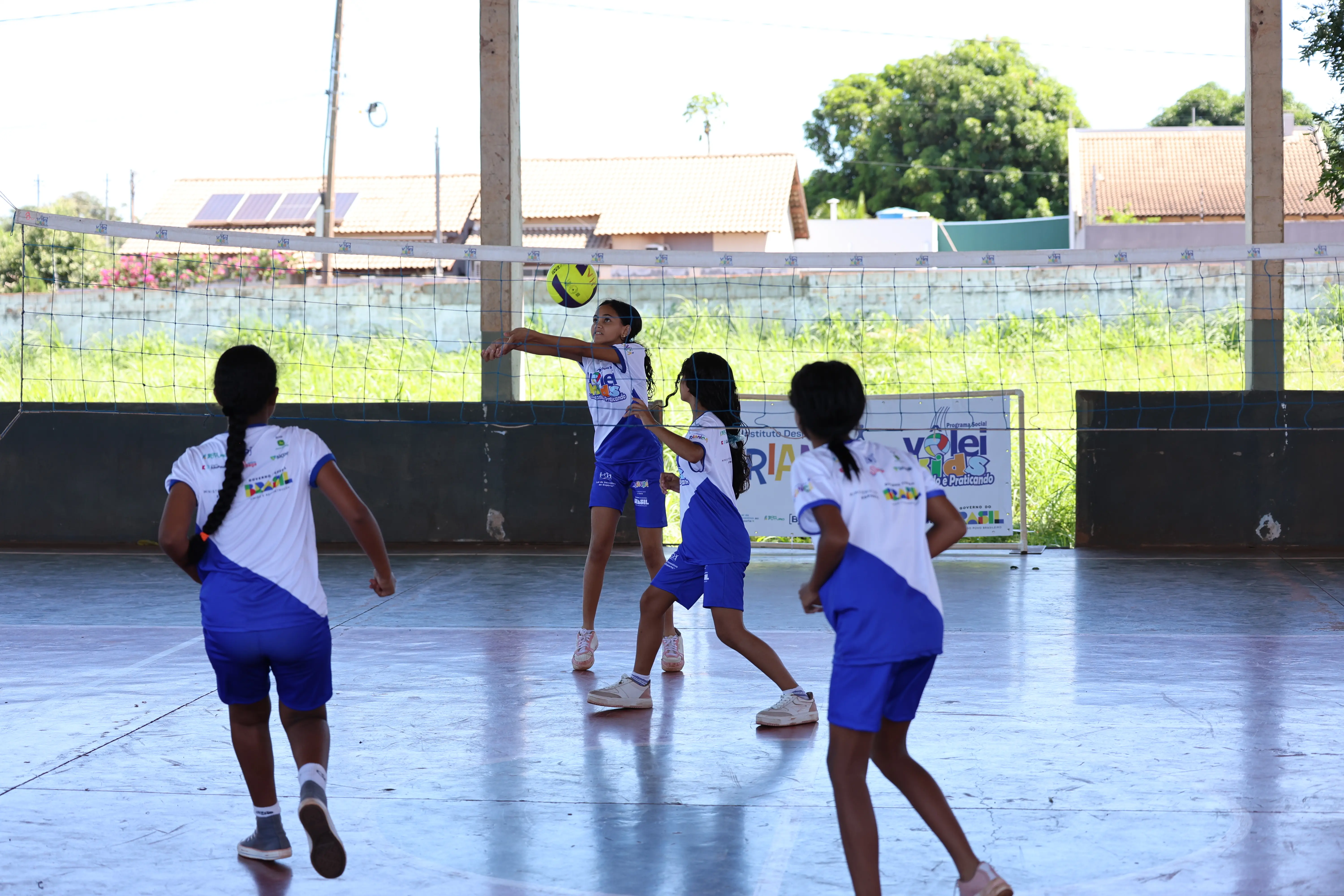 Grupo joga em quadra coberta, com crianças correndo atrás da bola. Espaço amplo e iluminado.