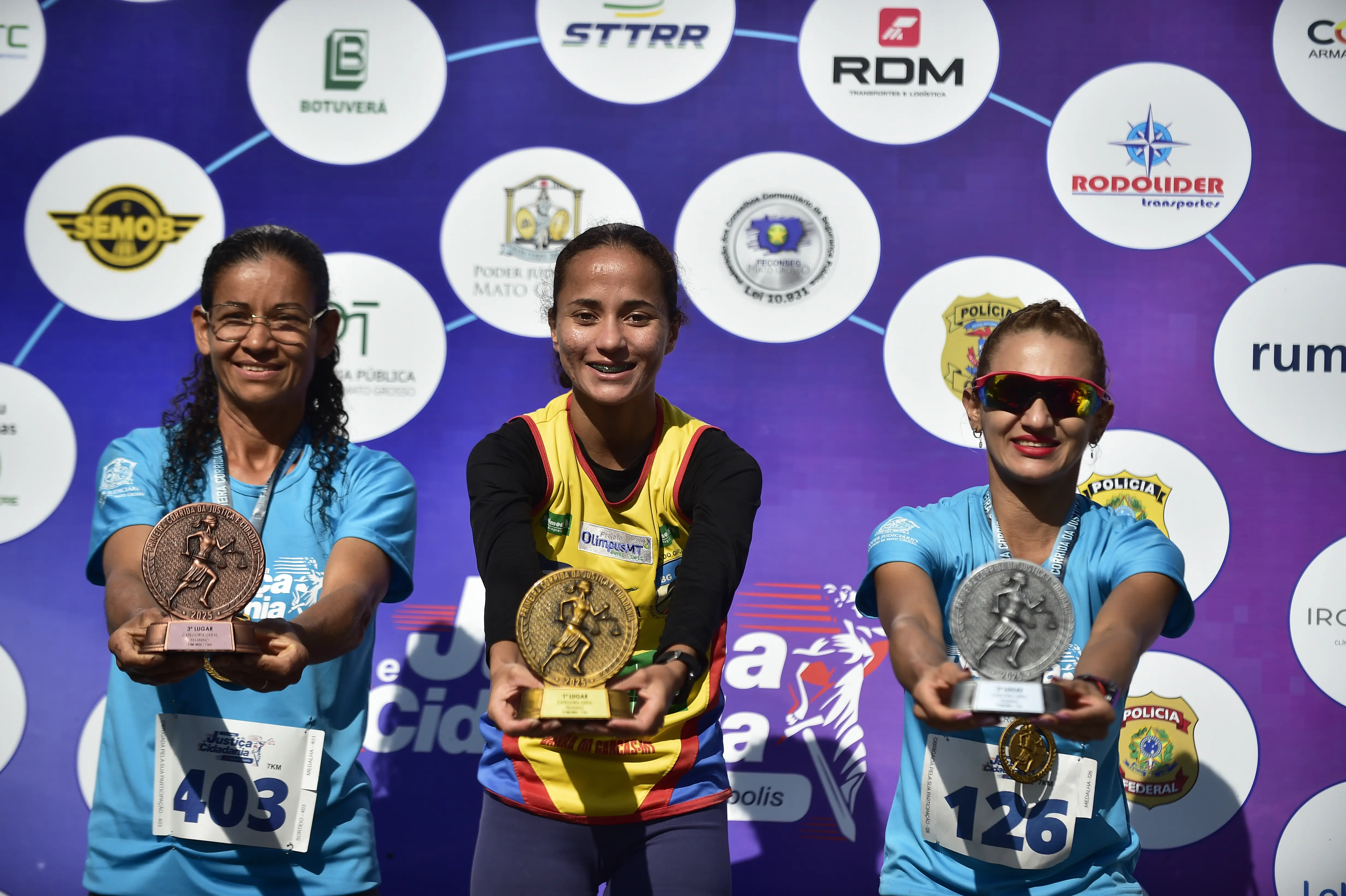 Três corredoras sorridentes no pódio exibem seus troféus de ouro, prata e bronze da Corrida da Justiça e Cidadania.