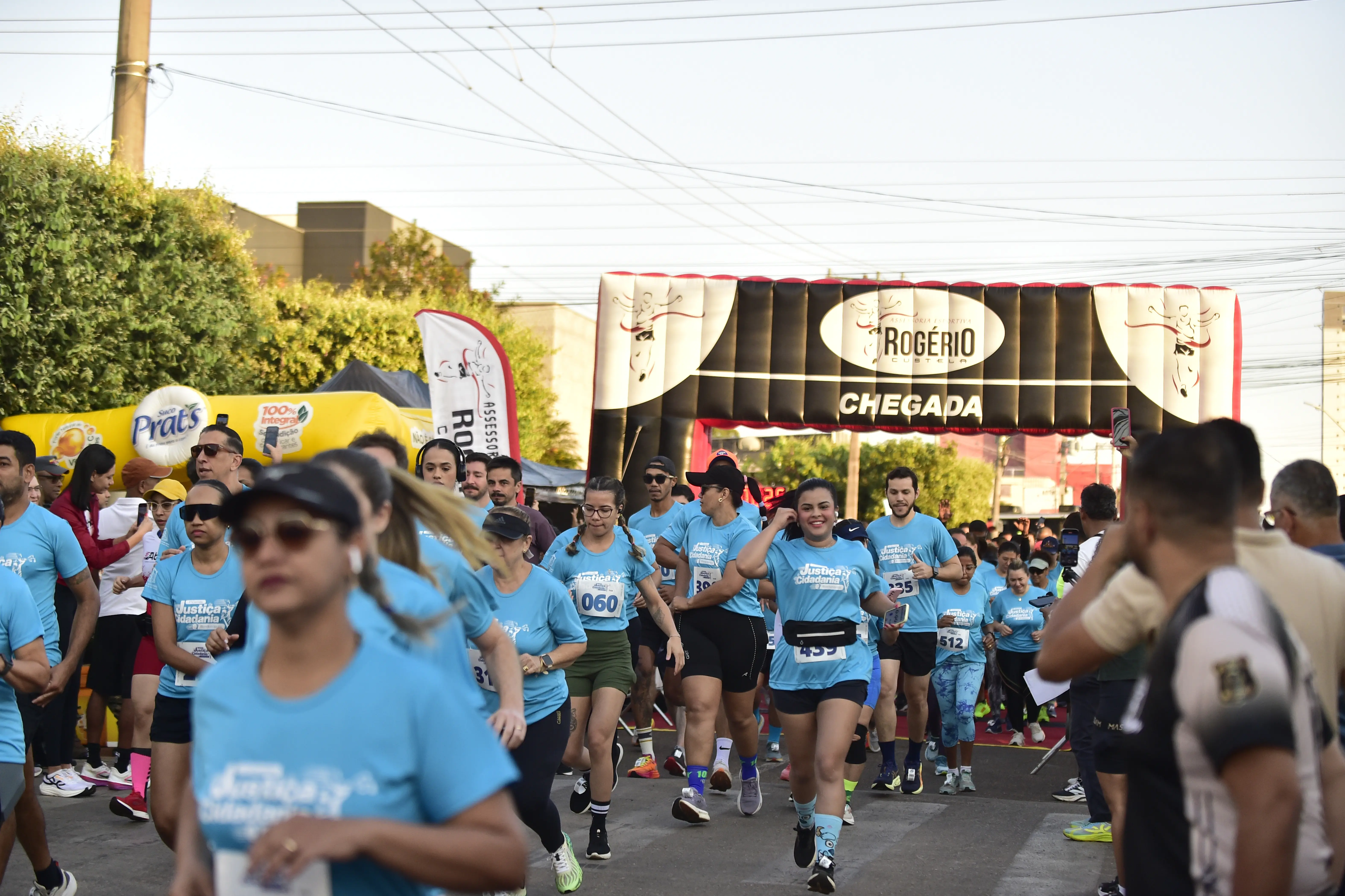 Dezenas de corredores em camisetas azuis cruzam a linha de chegada sob um arco inflável preto e vermelho.