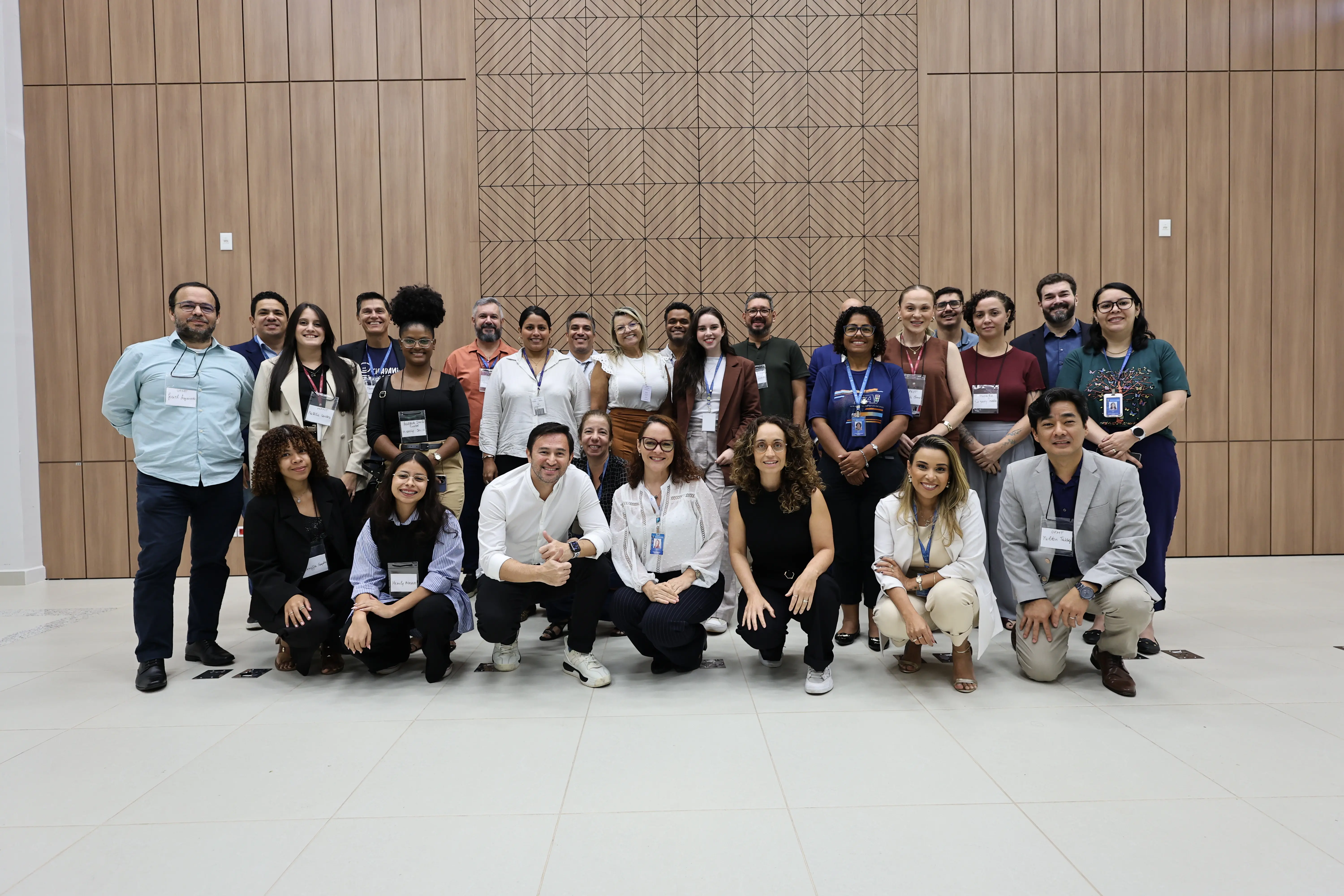 Foto em grupo com cerca de 25 pessoas posando sorridentes em um ambiente interno. Elas estão organizadas em duas fileiras, algumas agachadas, à frente de painéis de madeira.