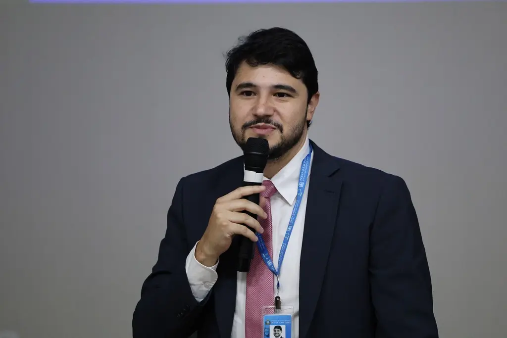 Foto horizontal em plano médio do juiz Victor Hugo Sousa Santos falando ao microfone, em pé. Ele é um jovem branco, de olhos, cabelo e barba pretos, usando camisa branca, gravata rosa e paletó azul marinho.